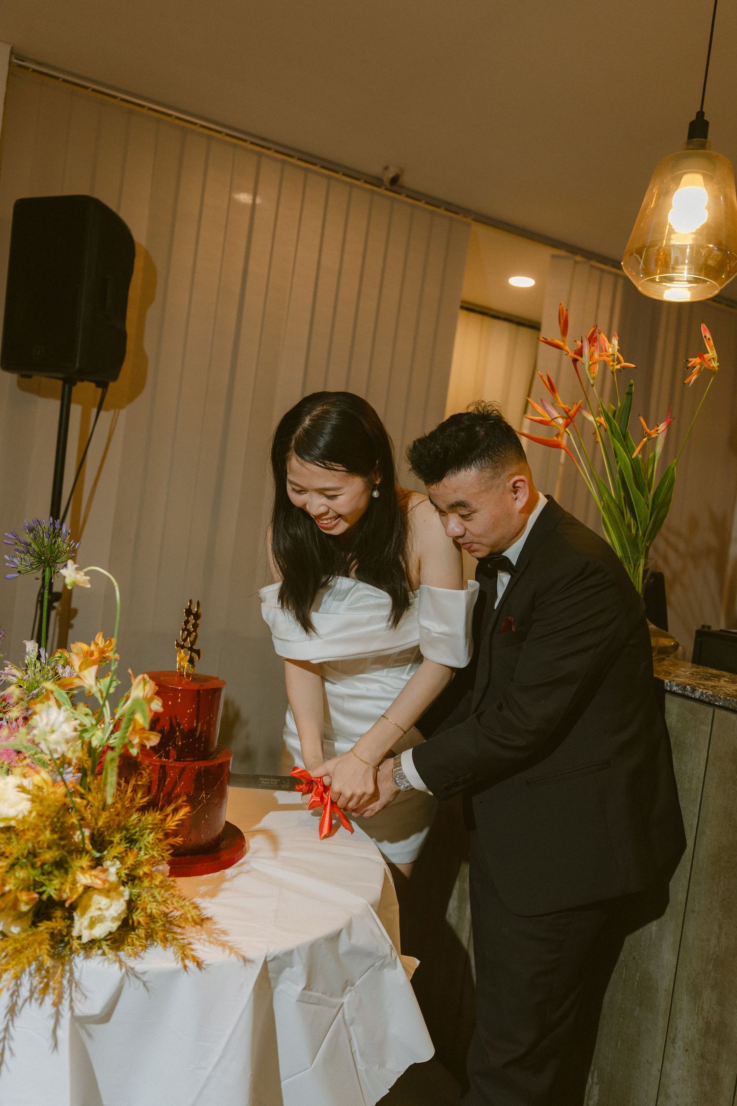 A couple celebrates at their wedding reception, performing traditional rituals with floral centerpieces on covered tables.