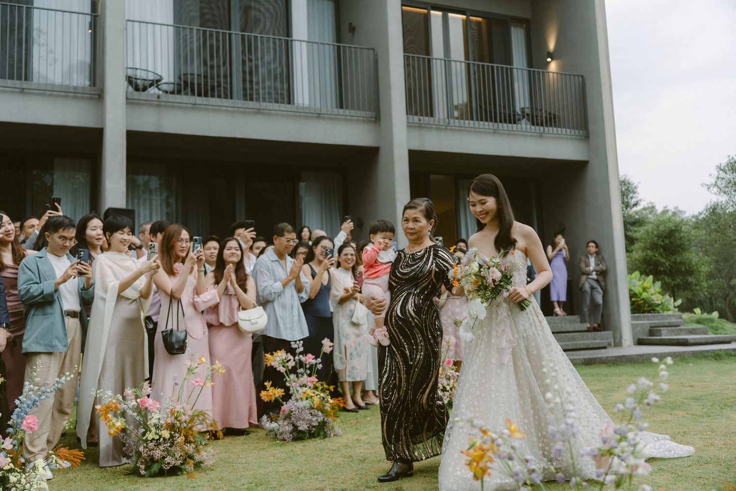 Wedding guests eagerly react during an outdoor celebration as a couple stands together holding bouquets.
