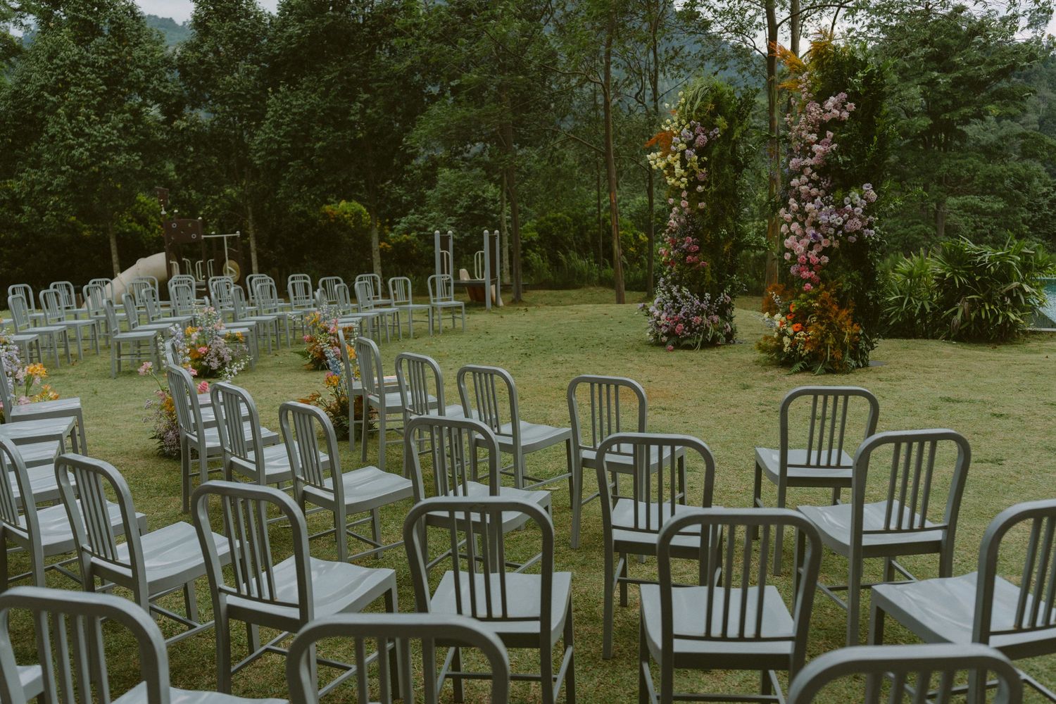 Rows of empty metal chairs arranged outdoors in a garden setting with lush greenery and flowers in the background.