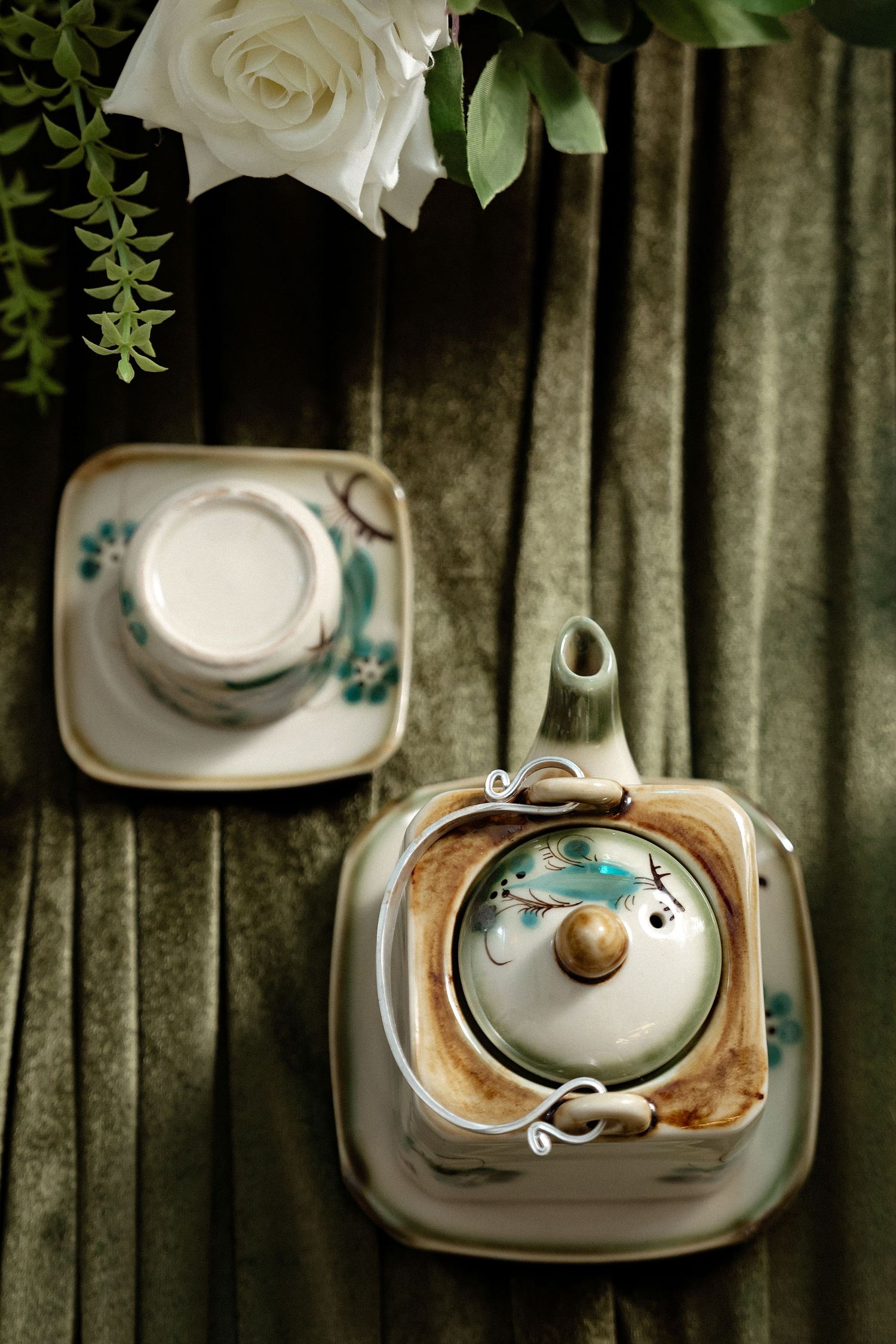 Overhead view of vintage teal floral teapot and cup set with white rose on wooden surface against dark curtain backdrop.