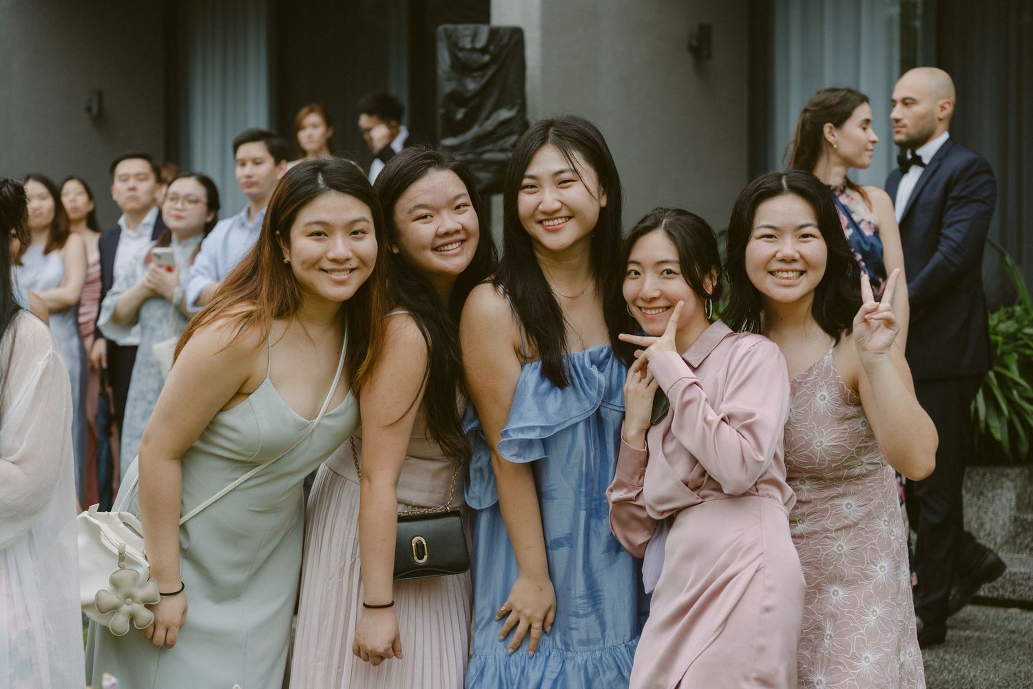 A group of friends in pastel dresses and casual attire pose together outdoors while smiling and making peace signs.