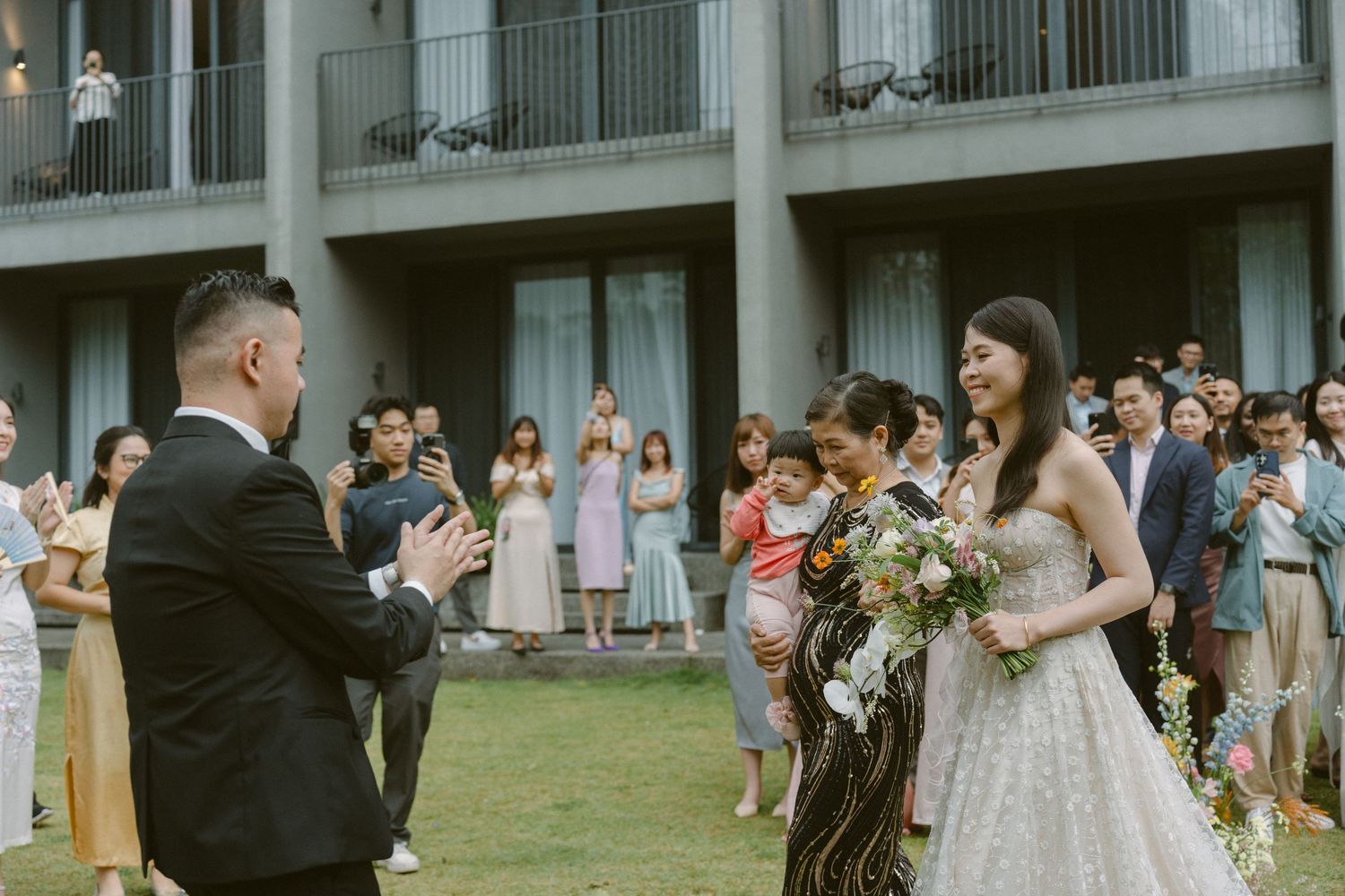 Outdoor wedding ceremony on a grassy lawn with guests gathered in front of a modern building with balconies.