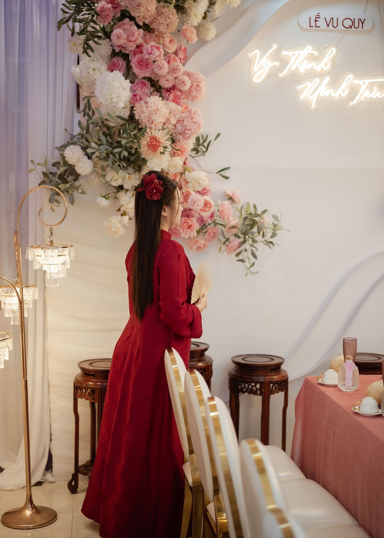 A person in a flowing red dress stands beside elegant floral arrangements and decorative tables at a formal event.