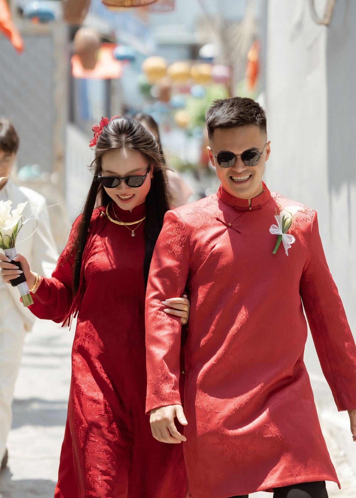 Two people in coordinating red traditional Chinese outfits walk arm in arm while wearing sunglasses on a sunny day.