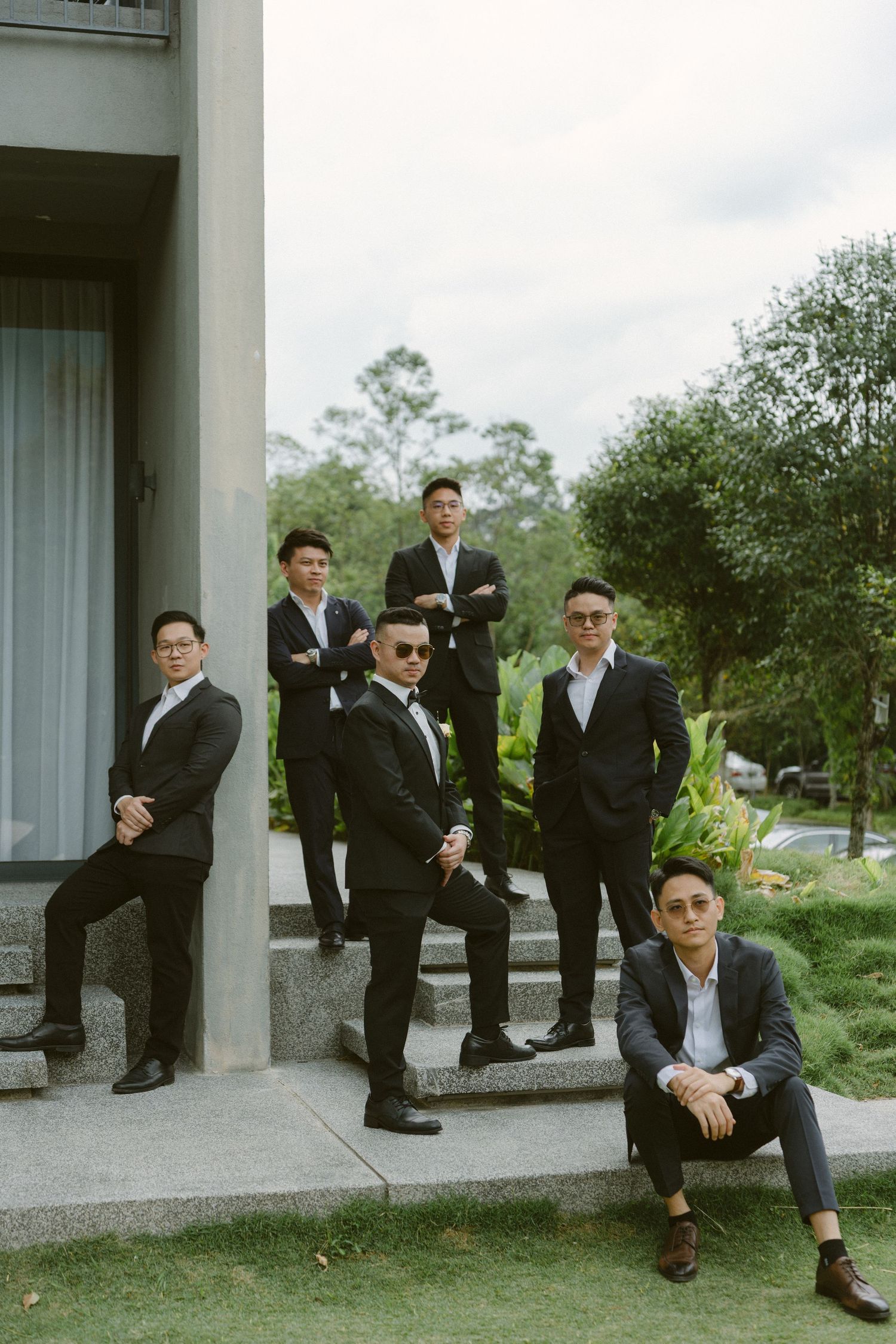 A group of men in black suits pose on outdoor concrete steps surrounded by greenery.