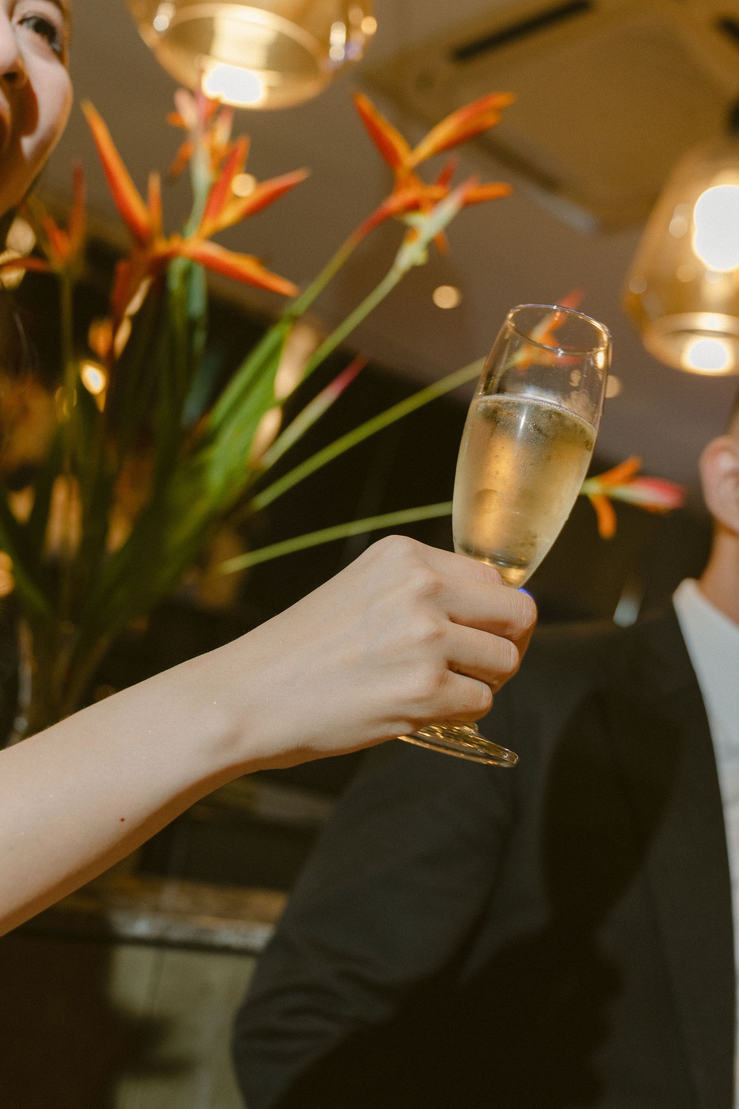 A hand holds up a champagne glass for a toast with floral decorations and warm lighting in the background.
