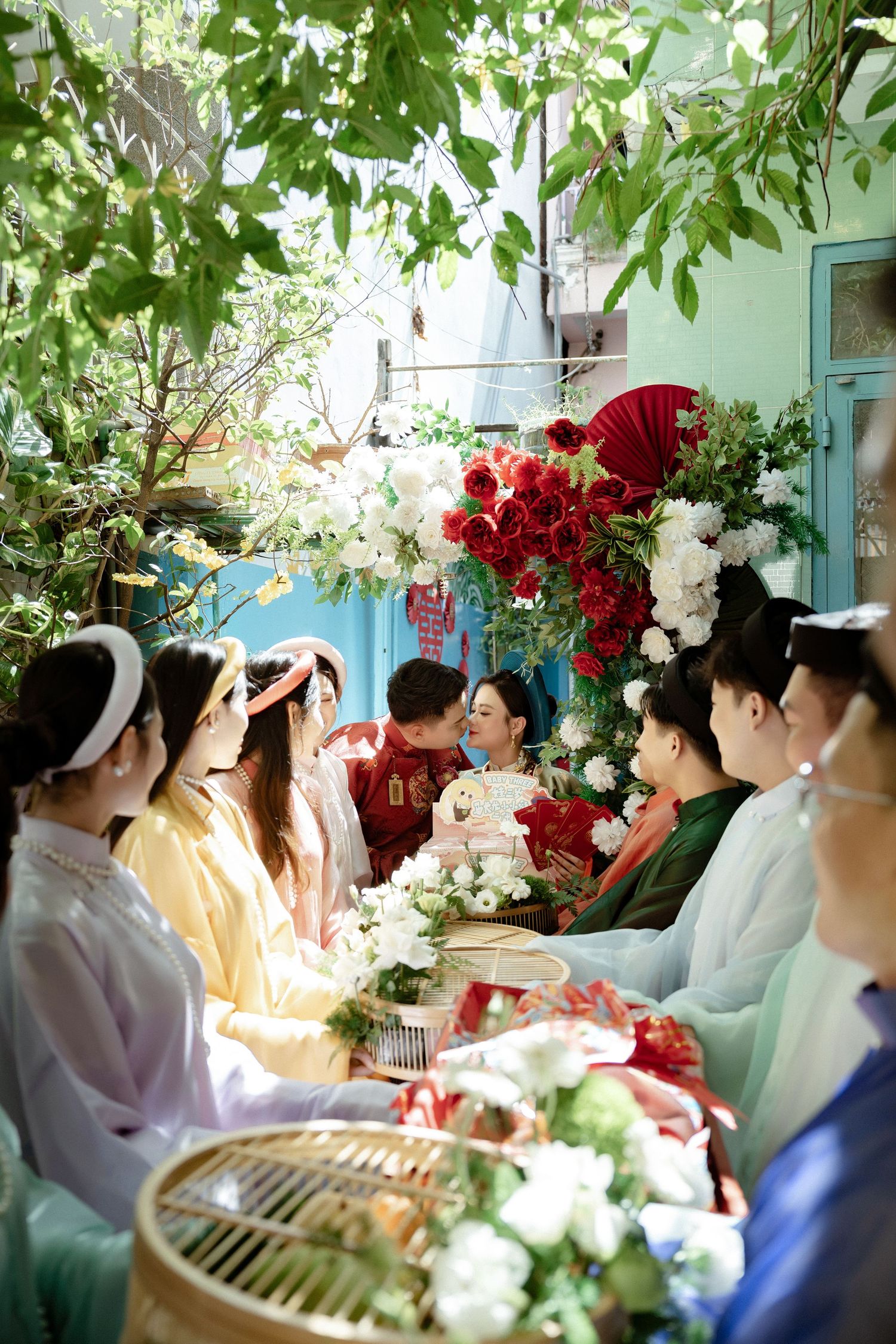 Traditional wedding ceremony with decorative red and white flowers and guests gathered under green foliage outdoors.
