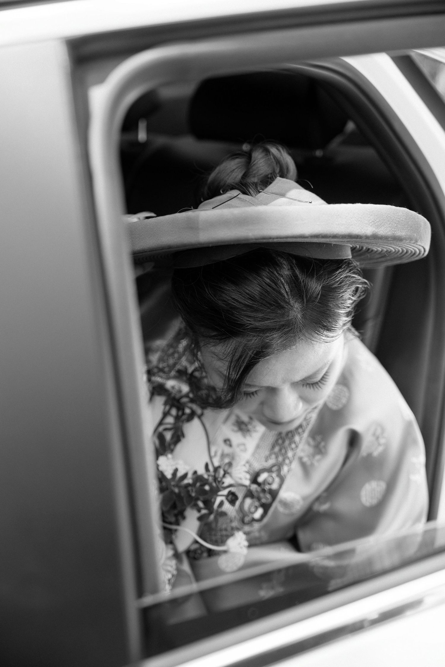 Black and white photograph showing a floral patterned dress and wide-brimmed hat viewed through a car window.