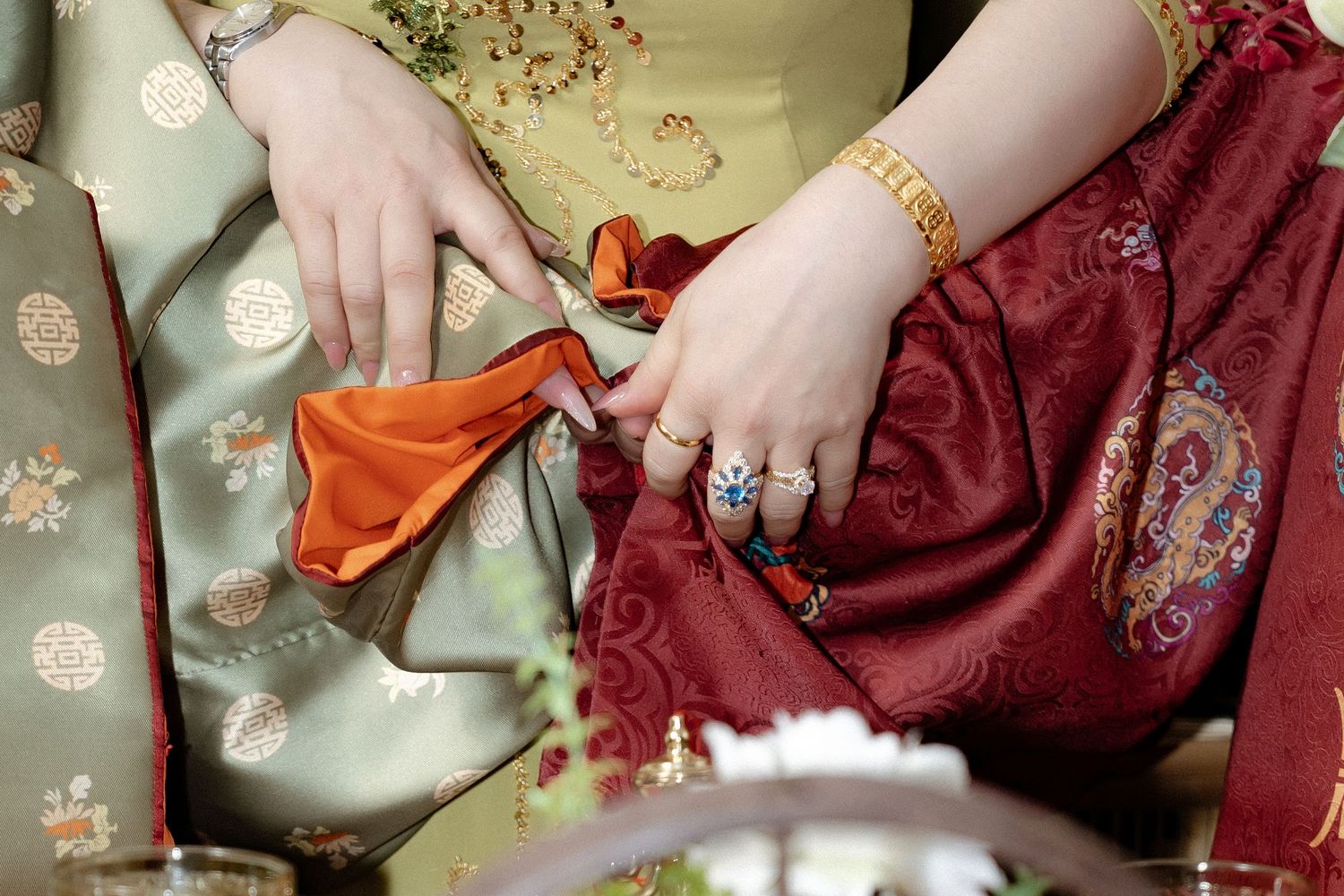 Hands wearing ornate rings rest on fabric with oriental patterns in sage green and burgundy silk.