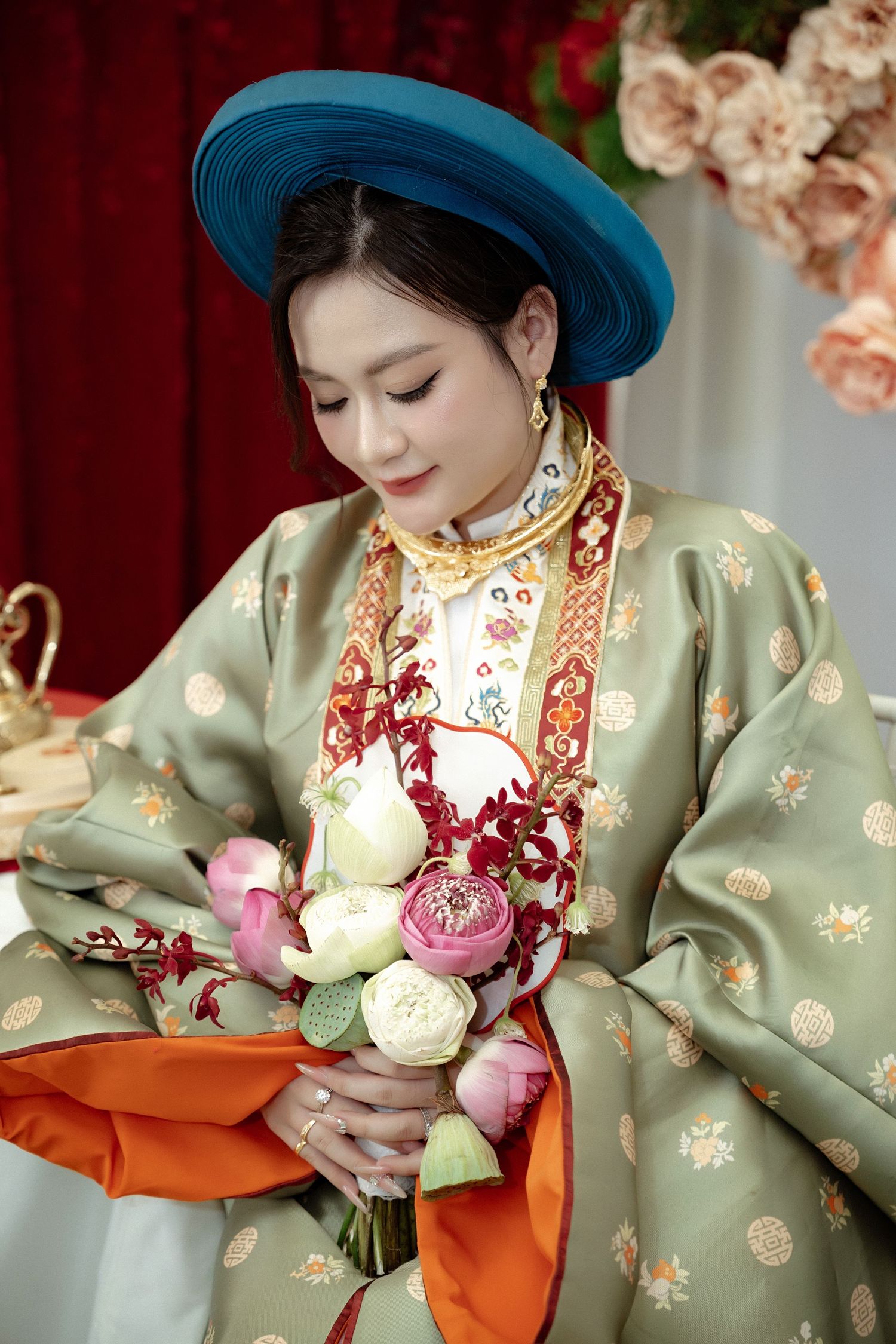 Traditional Vietnamese wedding attire with blue hat and sage green ao dai gown holding delicate pink and white floral bouquet.