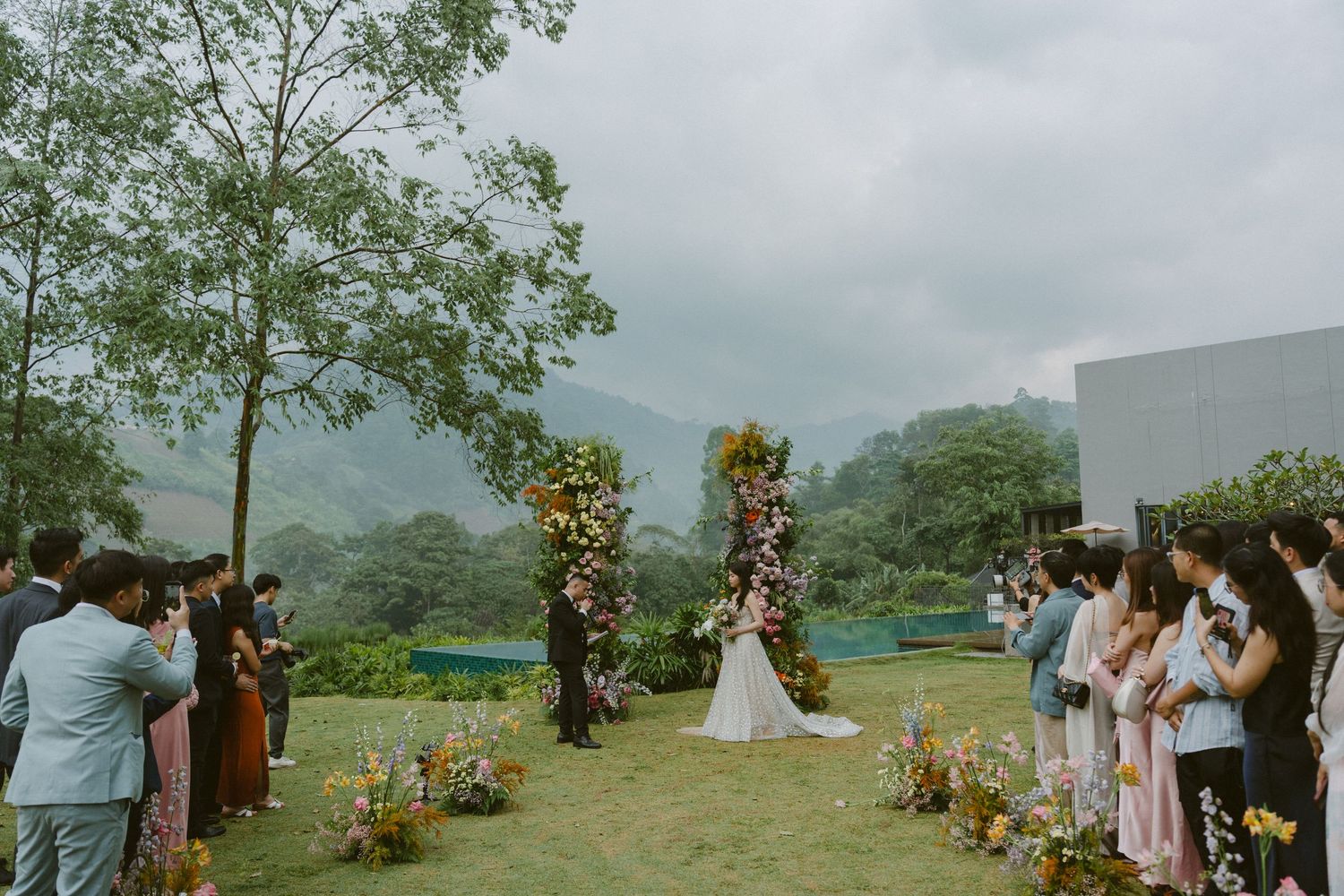 A wedding ceremony takes place on a misty hilltop with florally decorated arches and guests watching from both sides of the aisle.
