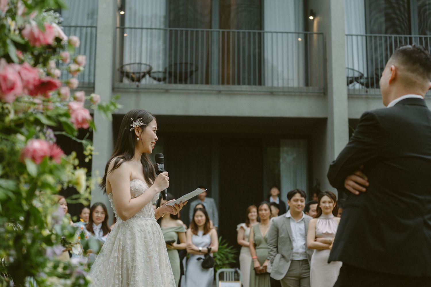 A bride recites vows during an outdoor garden wedding ceremony with pink flowers and guests in attendance.