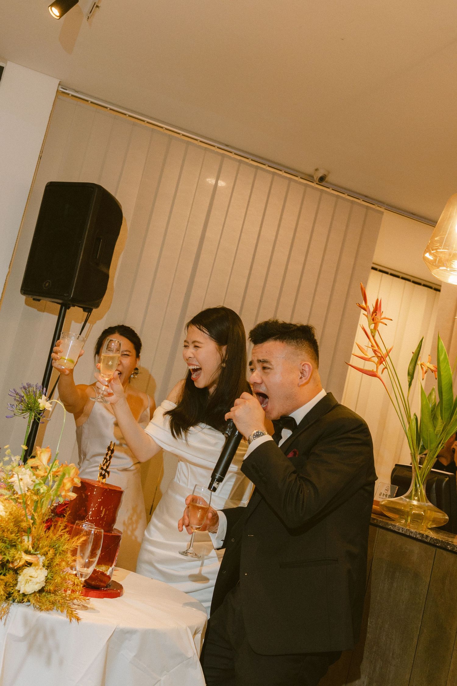 Indoor celebration event with people holding microphones and raising glasses in a decorated room with flowers.