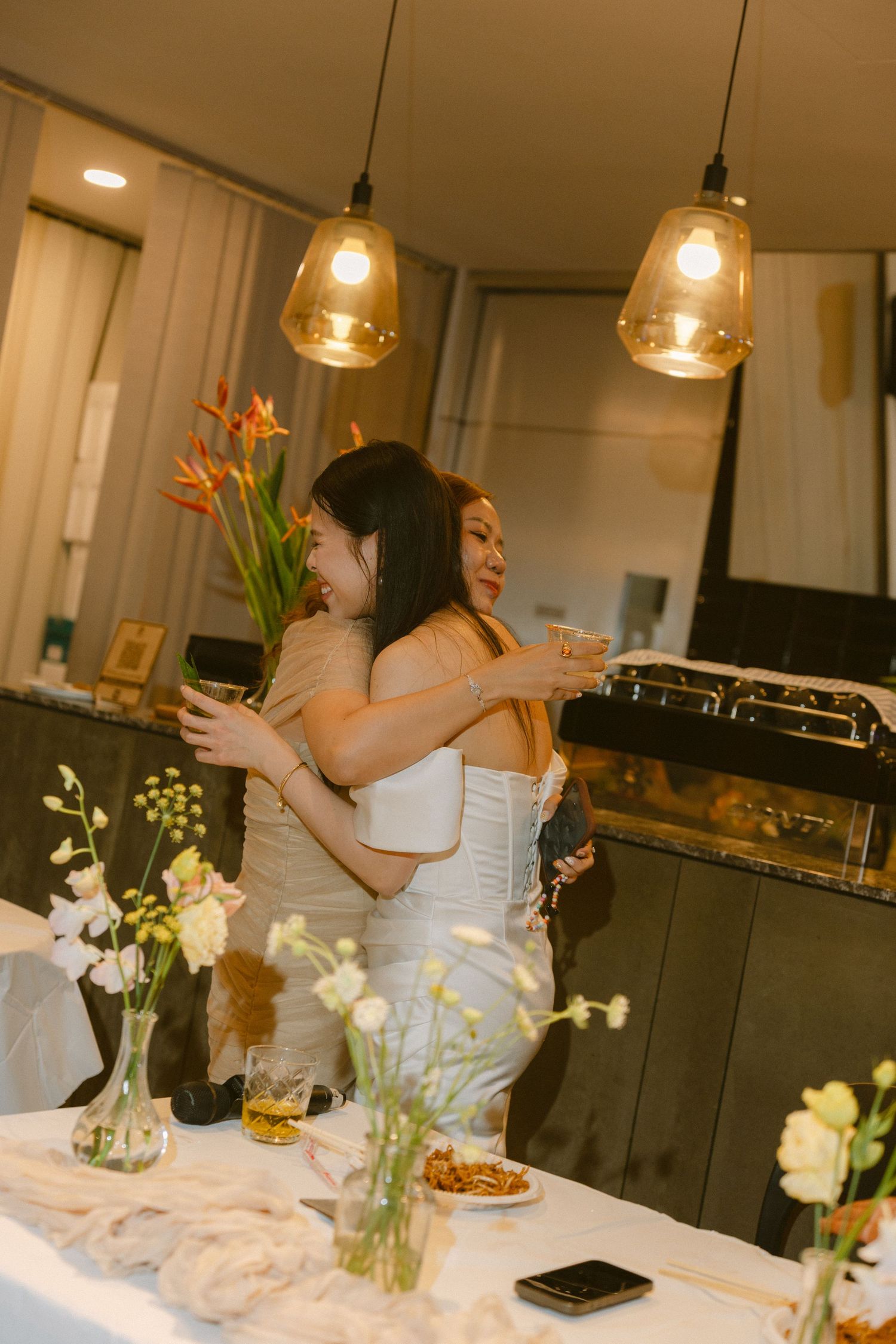 Two people in white dresses share an emotional embrace at an intimate dinner party decorated with floral centerpieces.