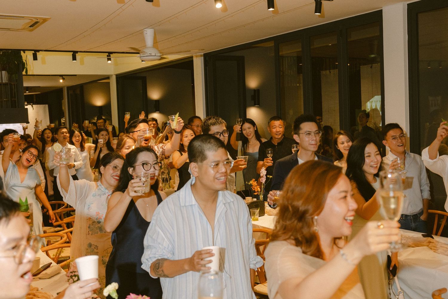 A crowded indoor event venue filled with guests in white attire celebrating at tables under warm lighting.