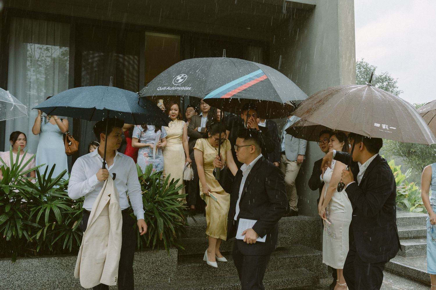 Wedding party holds umbrellas while gathering outside in the rain during a formal occasion.