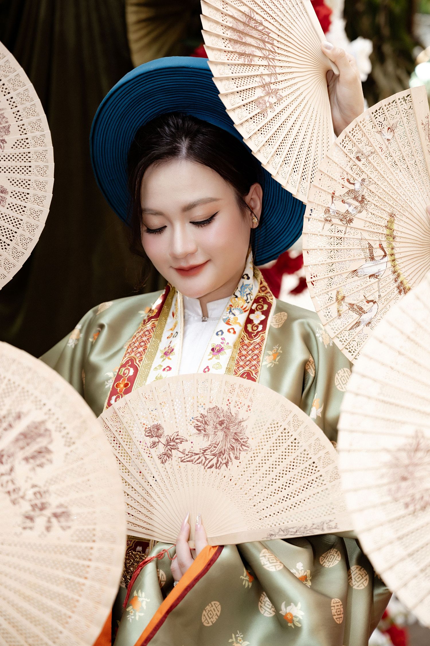 A person in traditional Asian attire poses with ornate white folding fans while wearing a blue hat and silk robe.