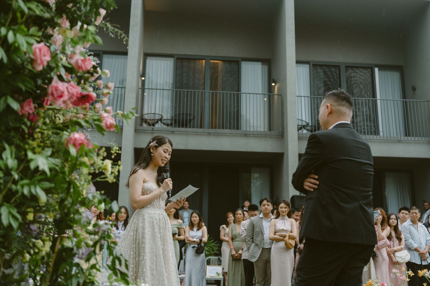 A wedding ceremony takes place on an outdoor patio surrounded by pink flowers and white columns.