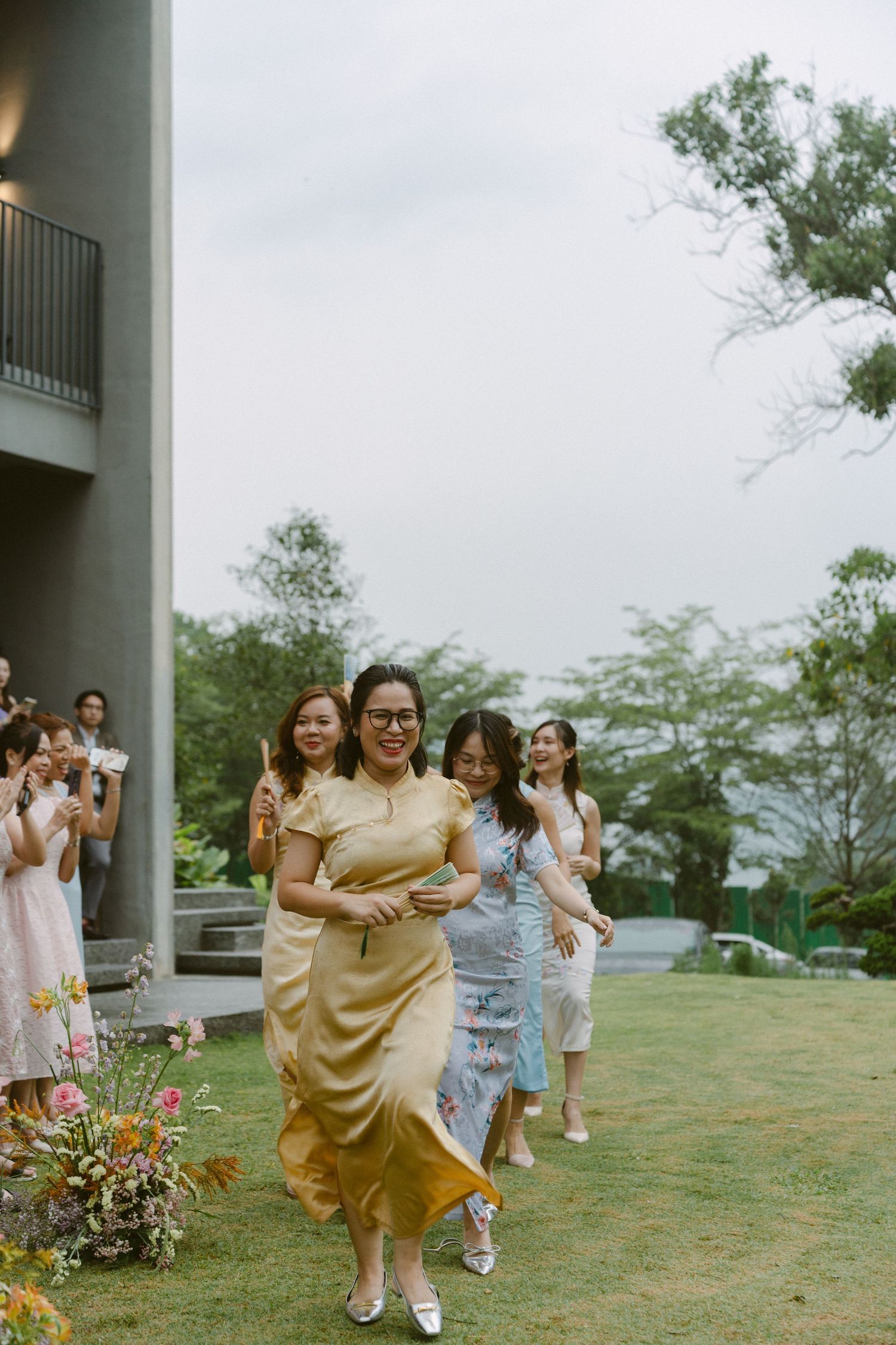 Women in elegant dresses run joyfully across a lawn during what appears to be a celebratory event outdoors.