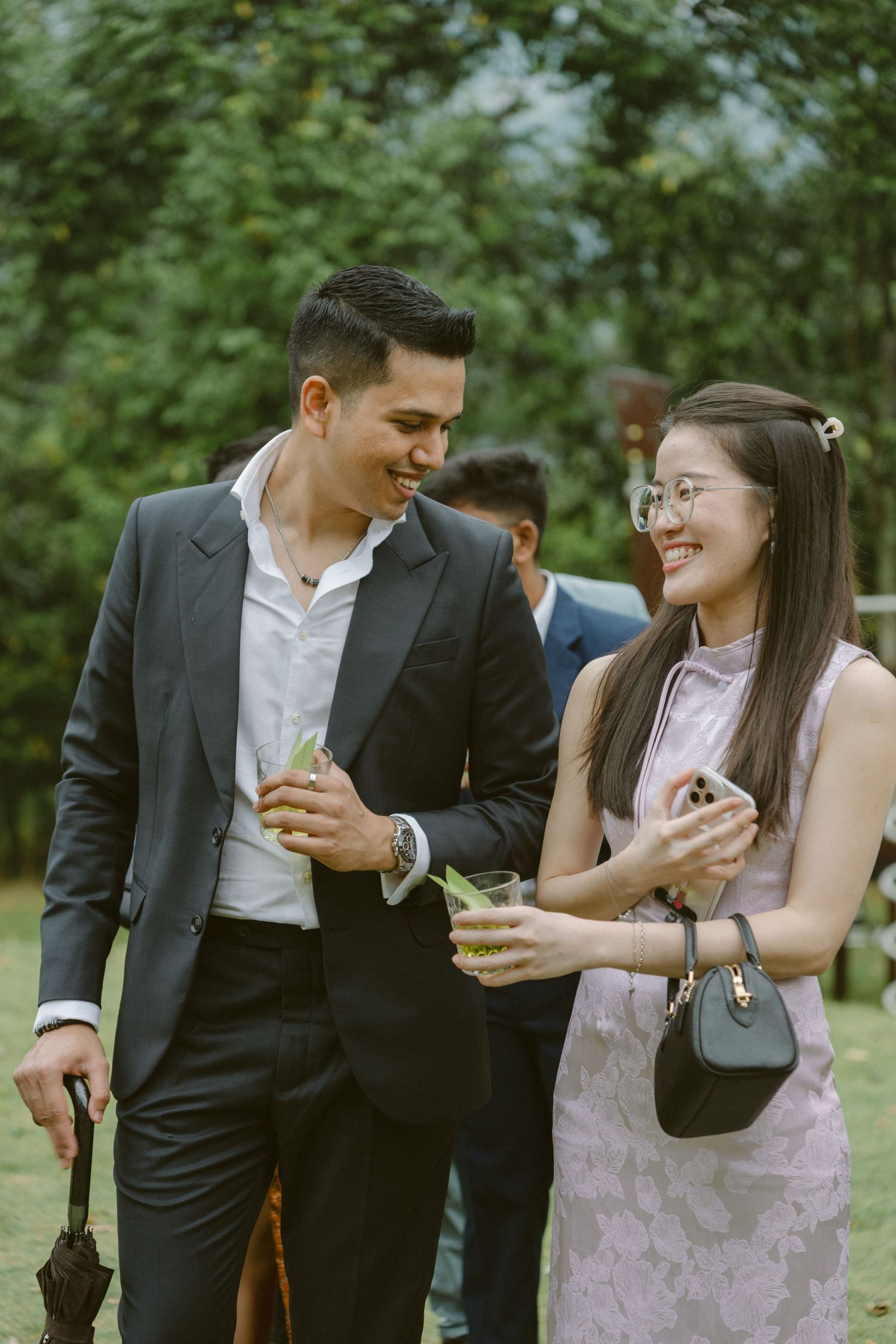 A couple in formal attire share joyful moments and laughter while enjoying drinks at an outdoor garden event.