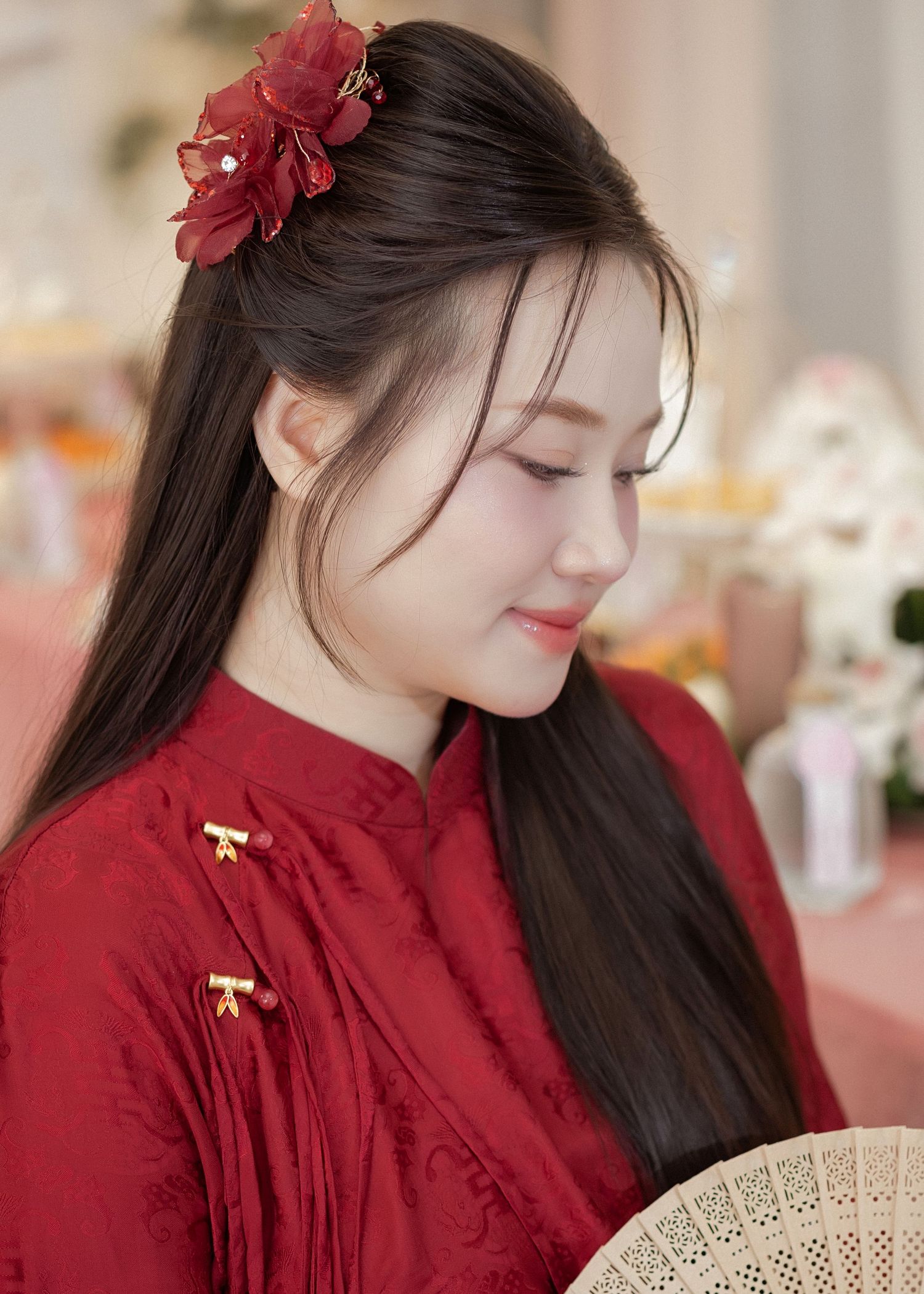 Person wearing a traditional red Chinese cheongsam dress and holding a decorative fan while looking down with a red flower in hair.