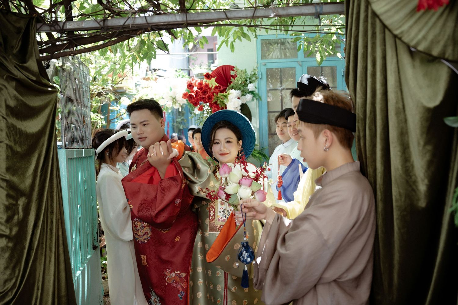 Three people in traditional Asian formal attire gather under decorative curtains at a cultural celebration ceremony.