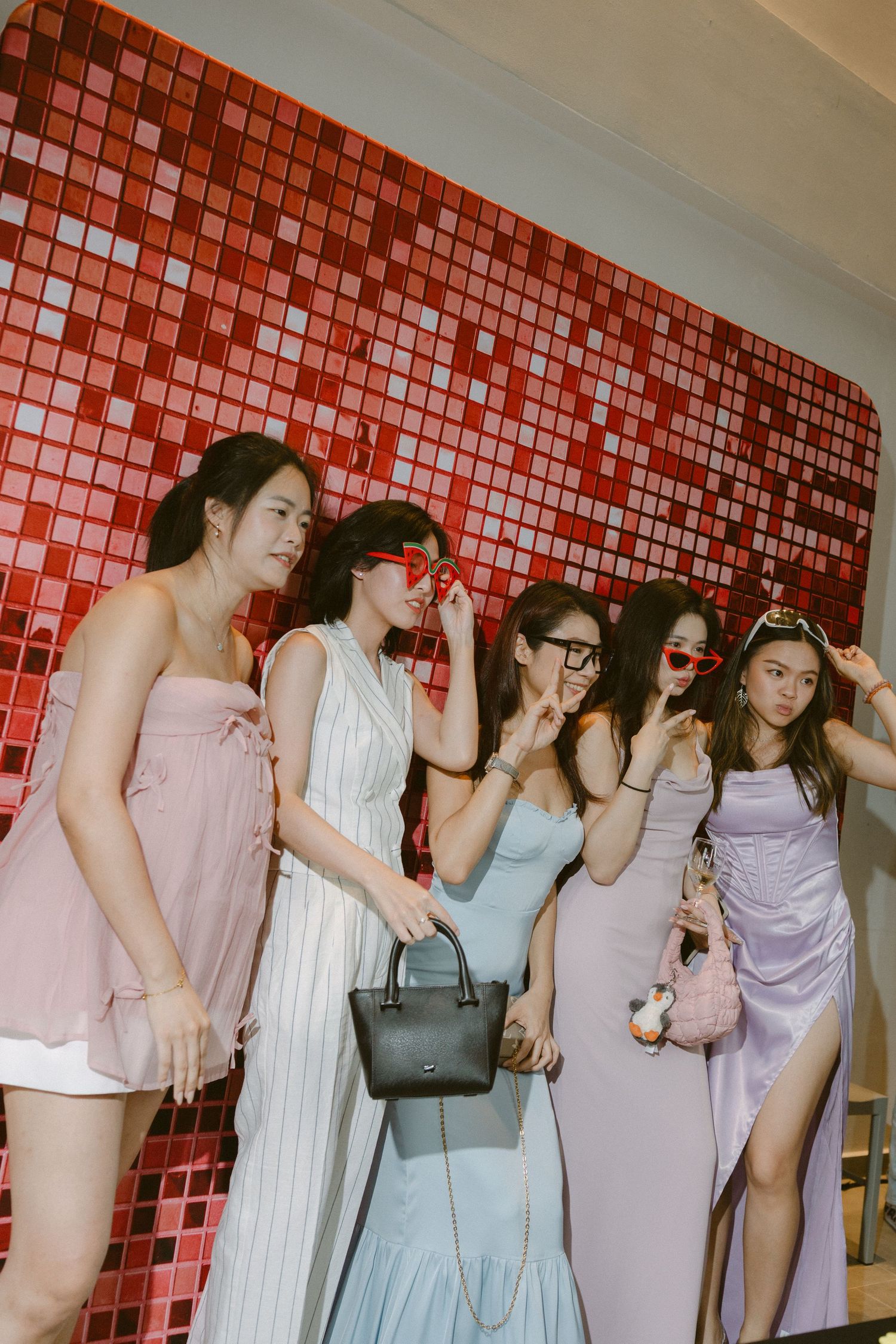 Group in pastel dresses posing together against a red mosaic tile wall in retro film style photography.