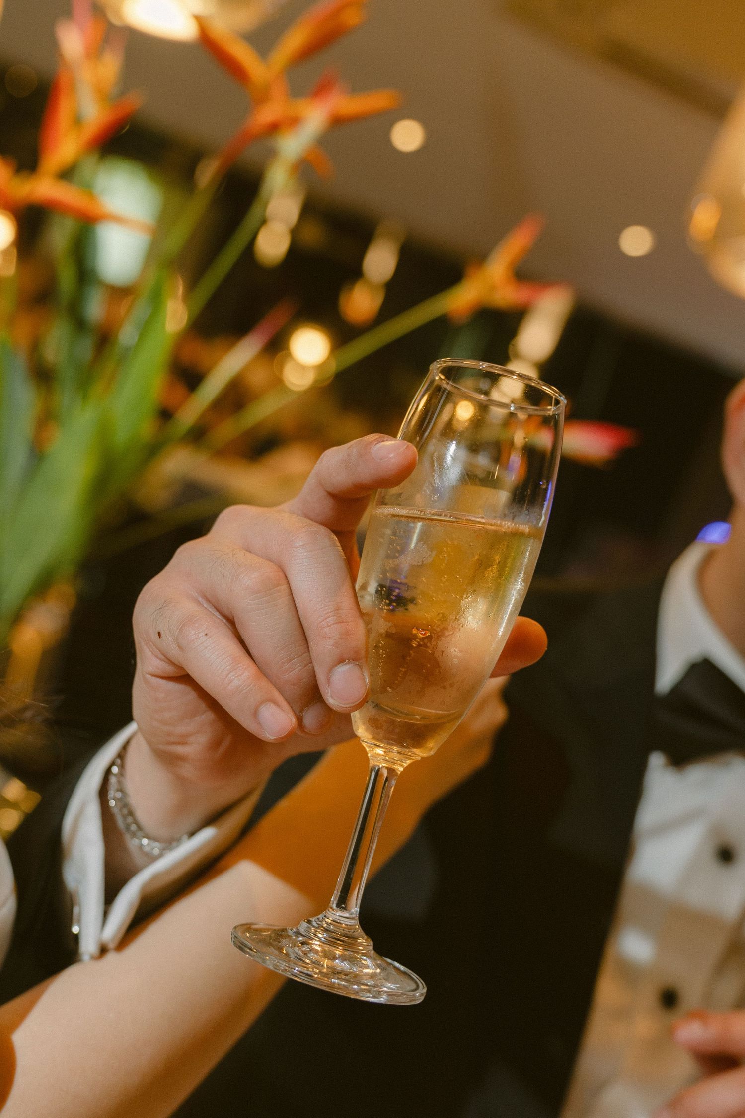 A hand holds up a champagne flute filled with bubbly golden liquid against a warm-lit background with orange flowers.