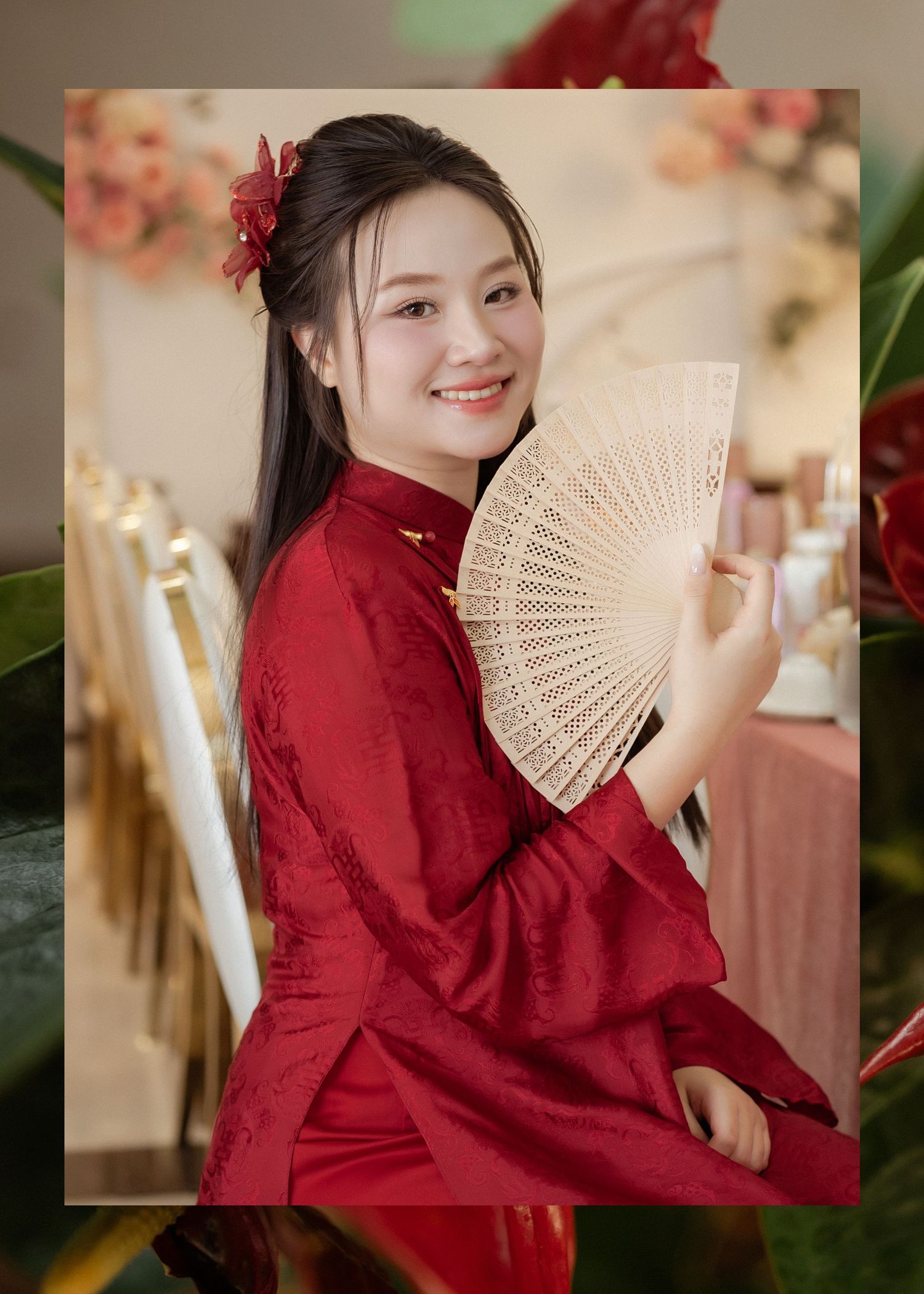 Traditional Asian woman in red cheongsam holding an ornate folding fan poses amid festive floral decorations.