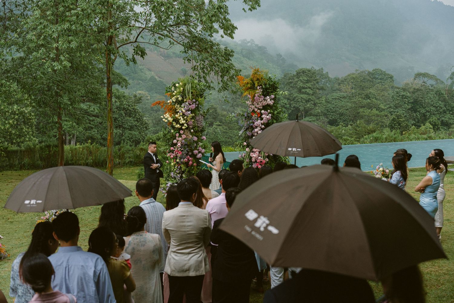 Wedding guests gather under umbrellas on a misty hillside during an outdoor ceremony with mountain views in background.