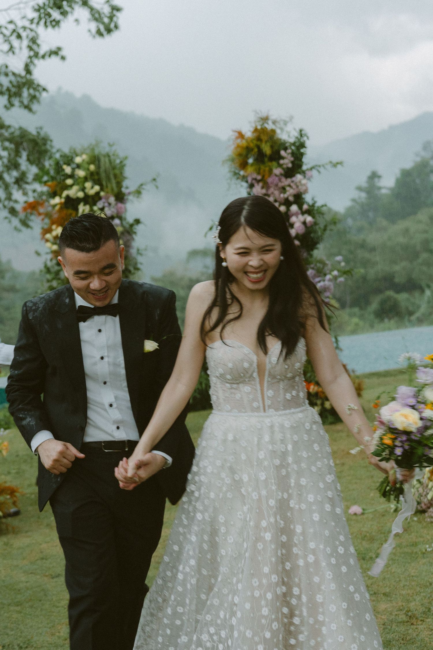A joyful outdoor wedding ceremony with a mountain backdrop as a couple walks together on lush grass with floral decorations.