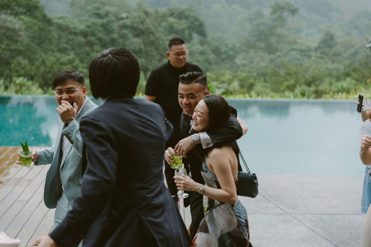 A group of people laughing and embracing on a wooden deck overlooking a serene turquoise lake surrounded by mountains.
