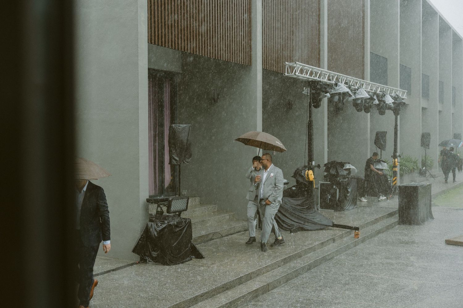 A person holding an umbrella walks down a rainy city street with blue-tinged lighting and buildings in the background.