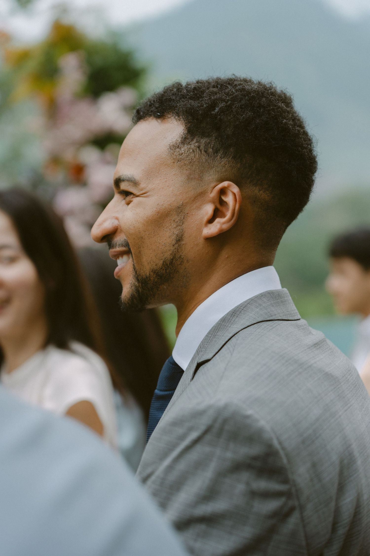 A guest in a gray suit laughs joyfully during an outdoor celebration with mountains visible in the background.