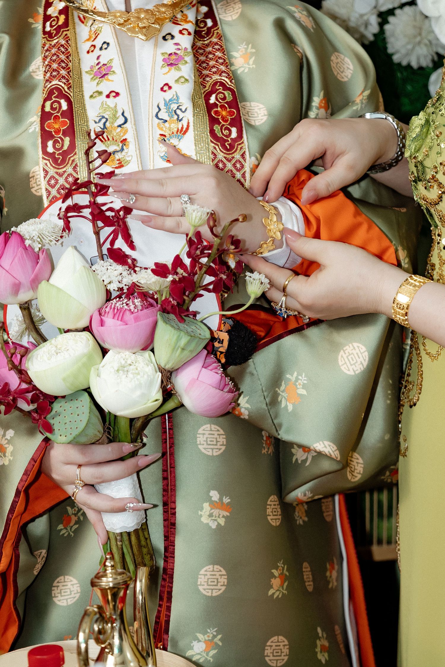 Hands holding vibrant pink and white lotus buds against sage green traditional Chinese silk robe with embroidered details.