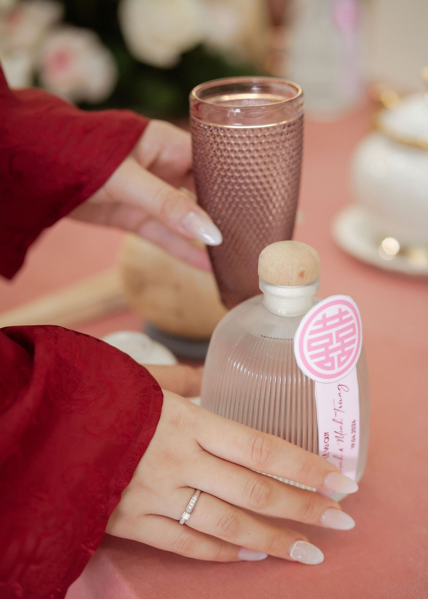 Hands with white manicure and a ring hold a decorative glass bottle and rose gold tumbler against a blurred background.