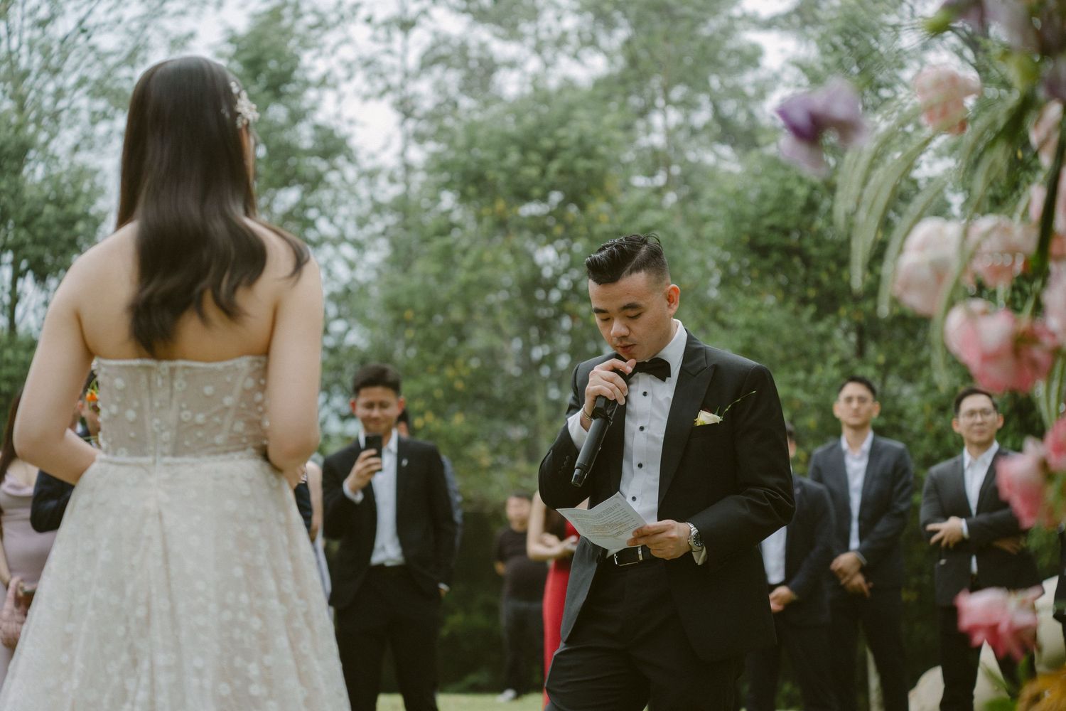 Bride and groom participate in a wedding ceremony on a lush green lawn surrounded by wedding party members.