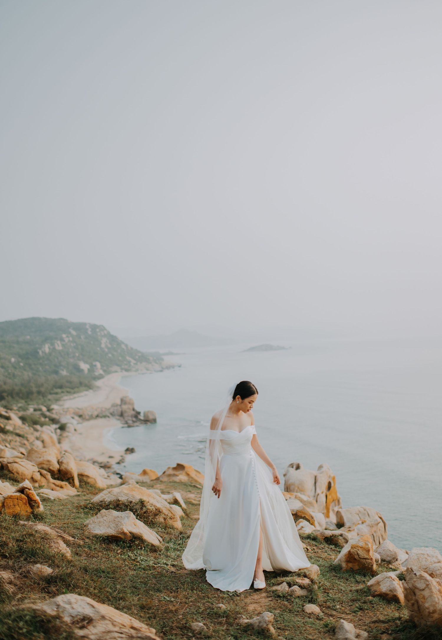 A bride in a flowing white dress stands on a rocky coastal cliff overlooking the ocean.