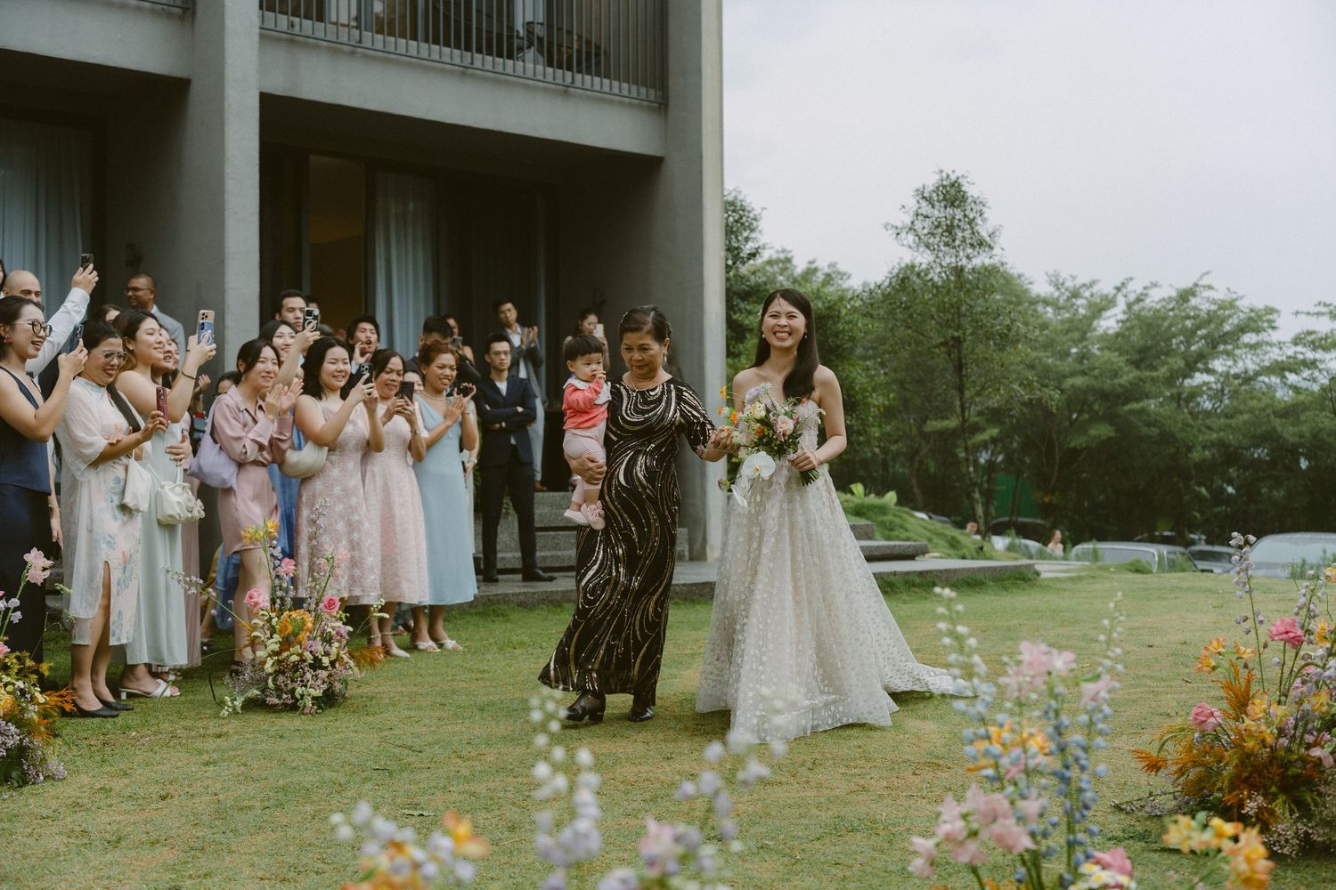 Wedding guests gather outdoors on a lawn with colorful wildflowers as a walk down the aisle moment unfolds.
