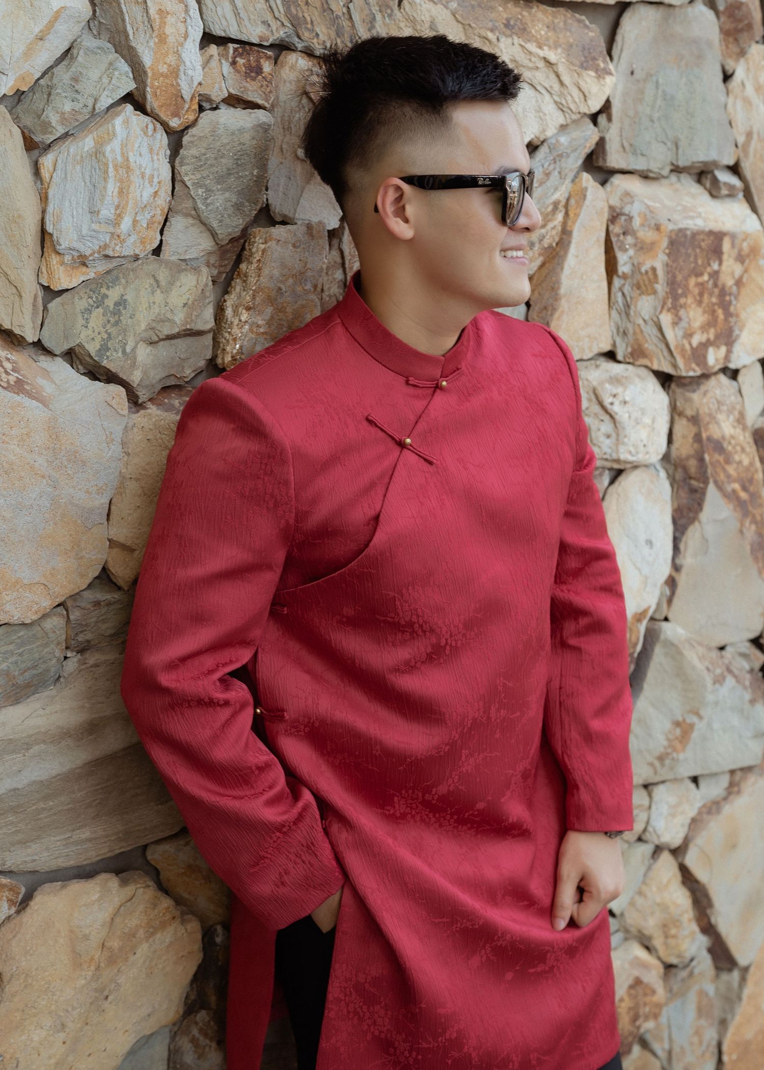 Portrait of someone wearing a red Chinese-style mandarin collar shirt and sunglasses against a stone wall background.