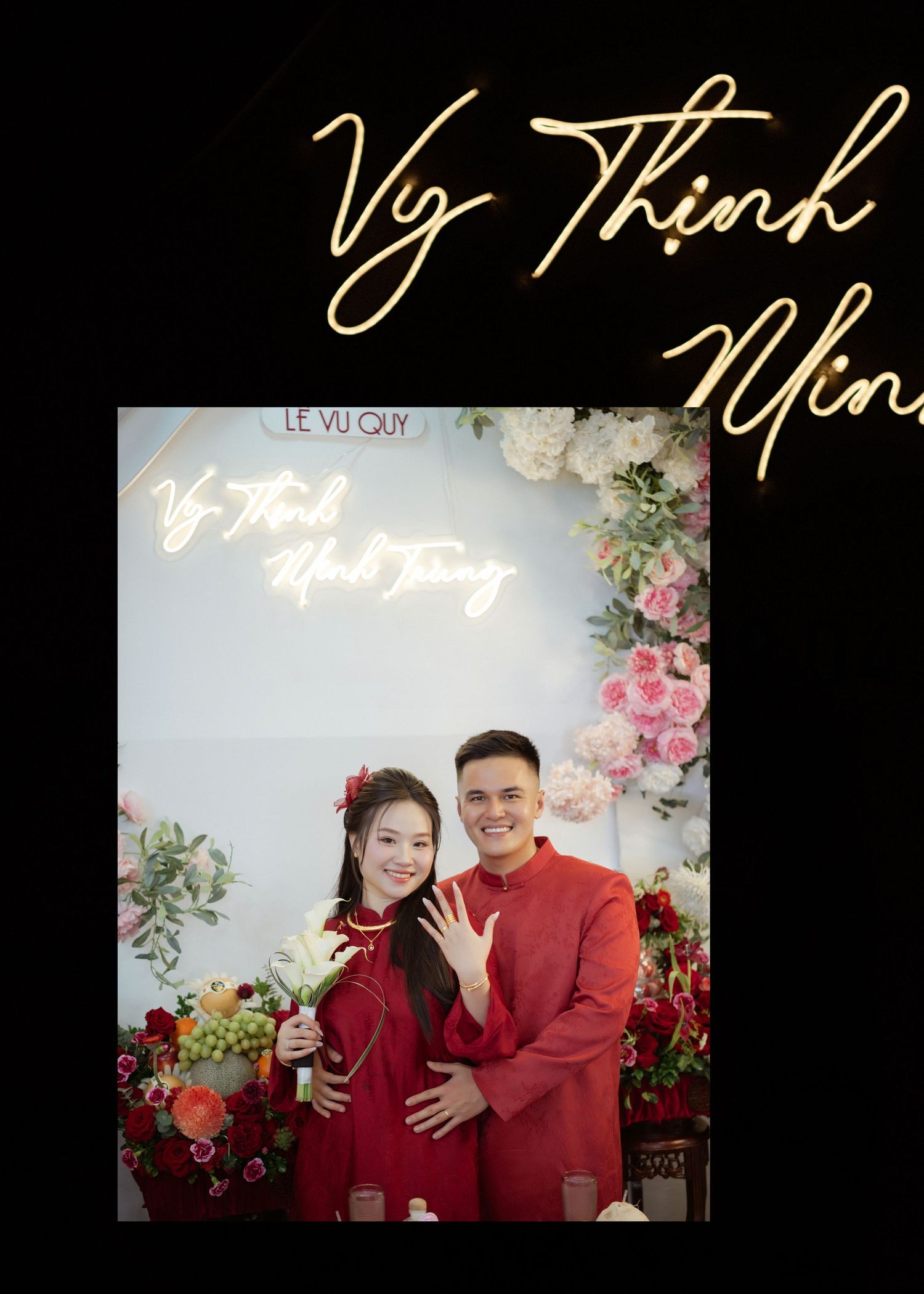 Elegant couple in matching red traditional outfits pose against floral backdrop with neon signage and pink roses.