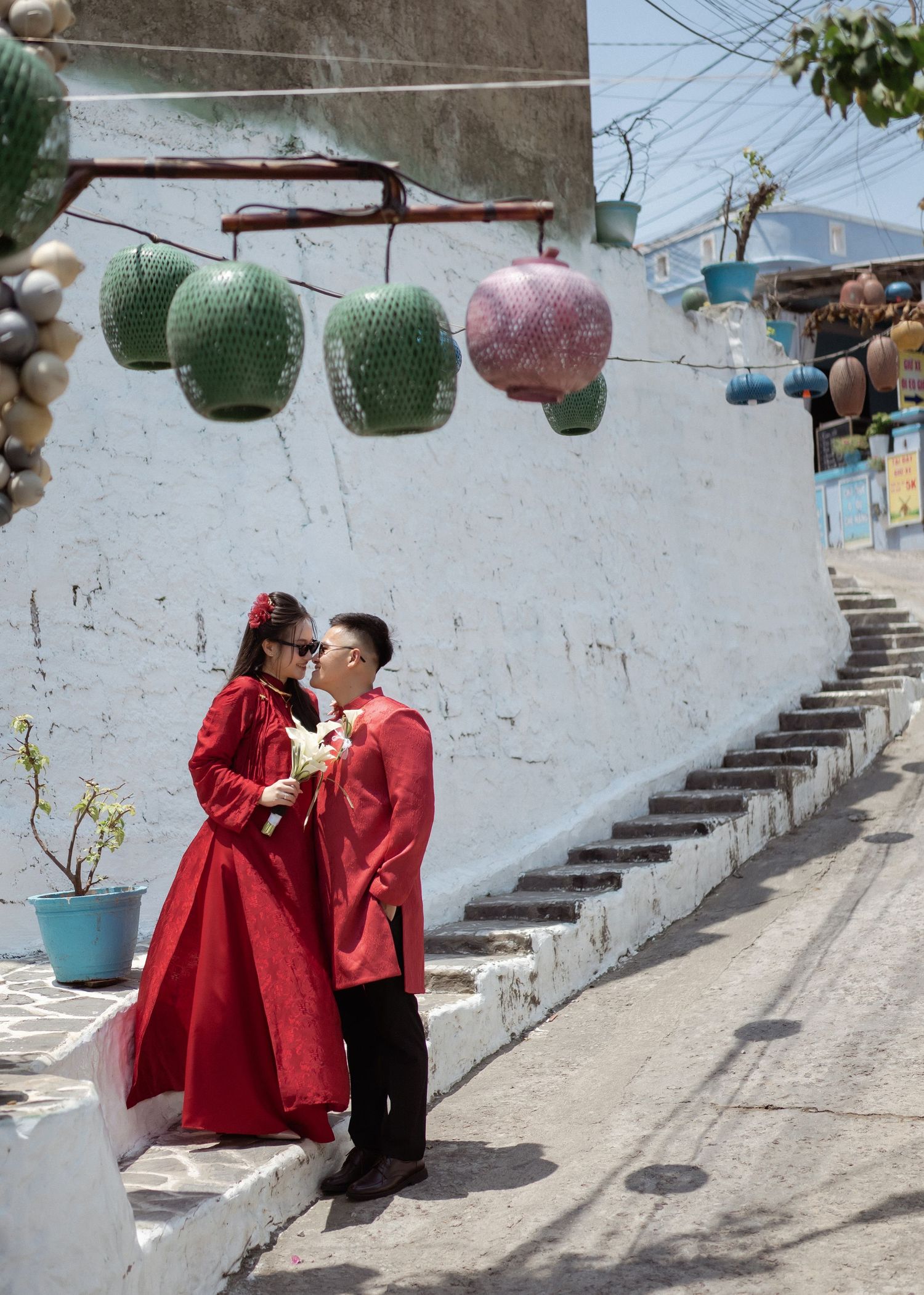 Two figures in matching red traditional outfits stand intimately on steps under decorative hanging lanterns against a white wall.