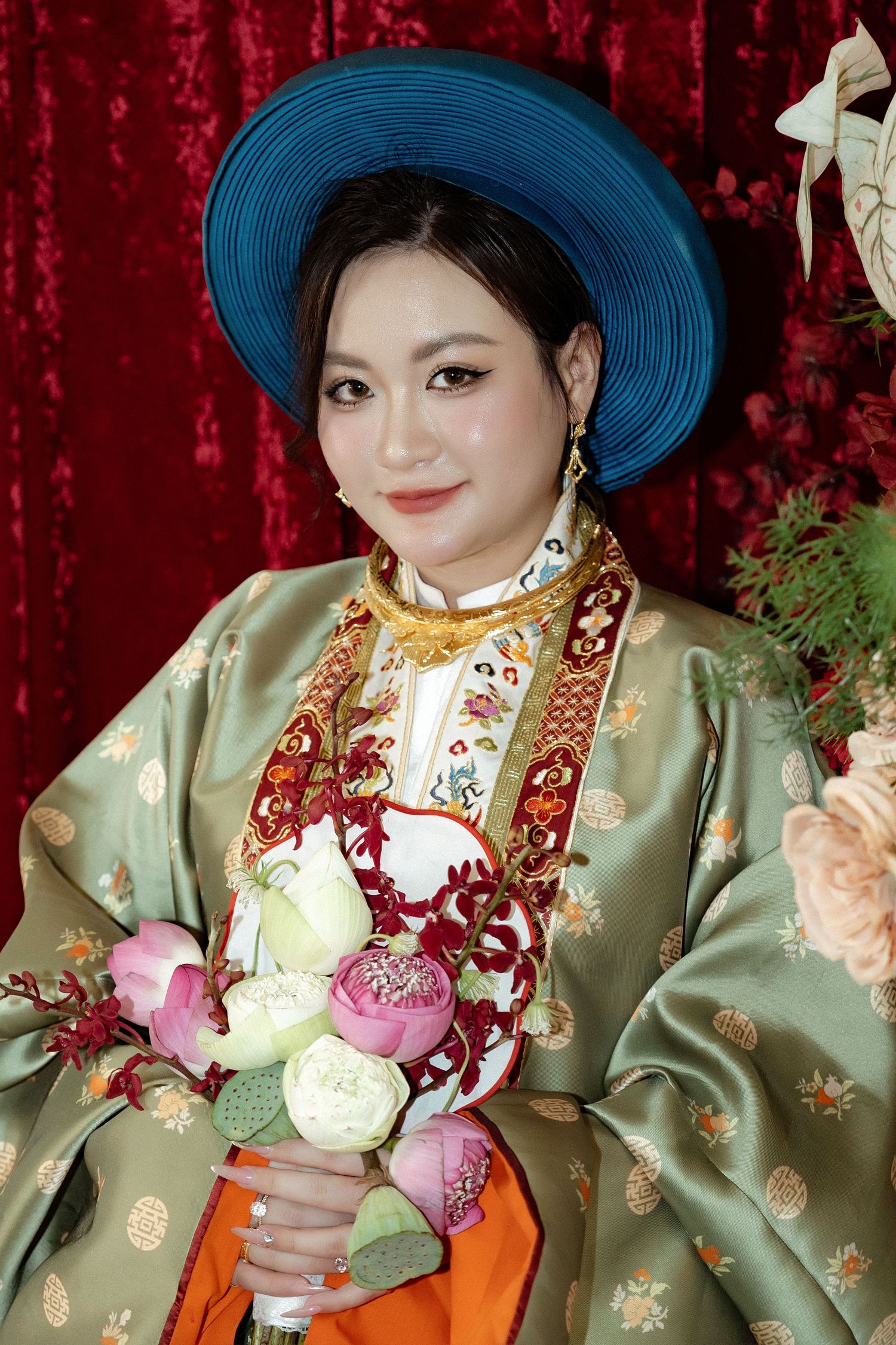 Person in traditional Vietnamese formal attire with blue hat and silk sage green ao dai holds pink and white flower bouquet.