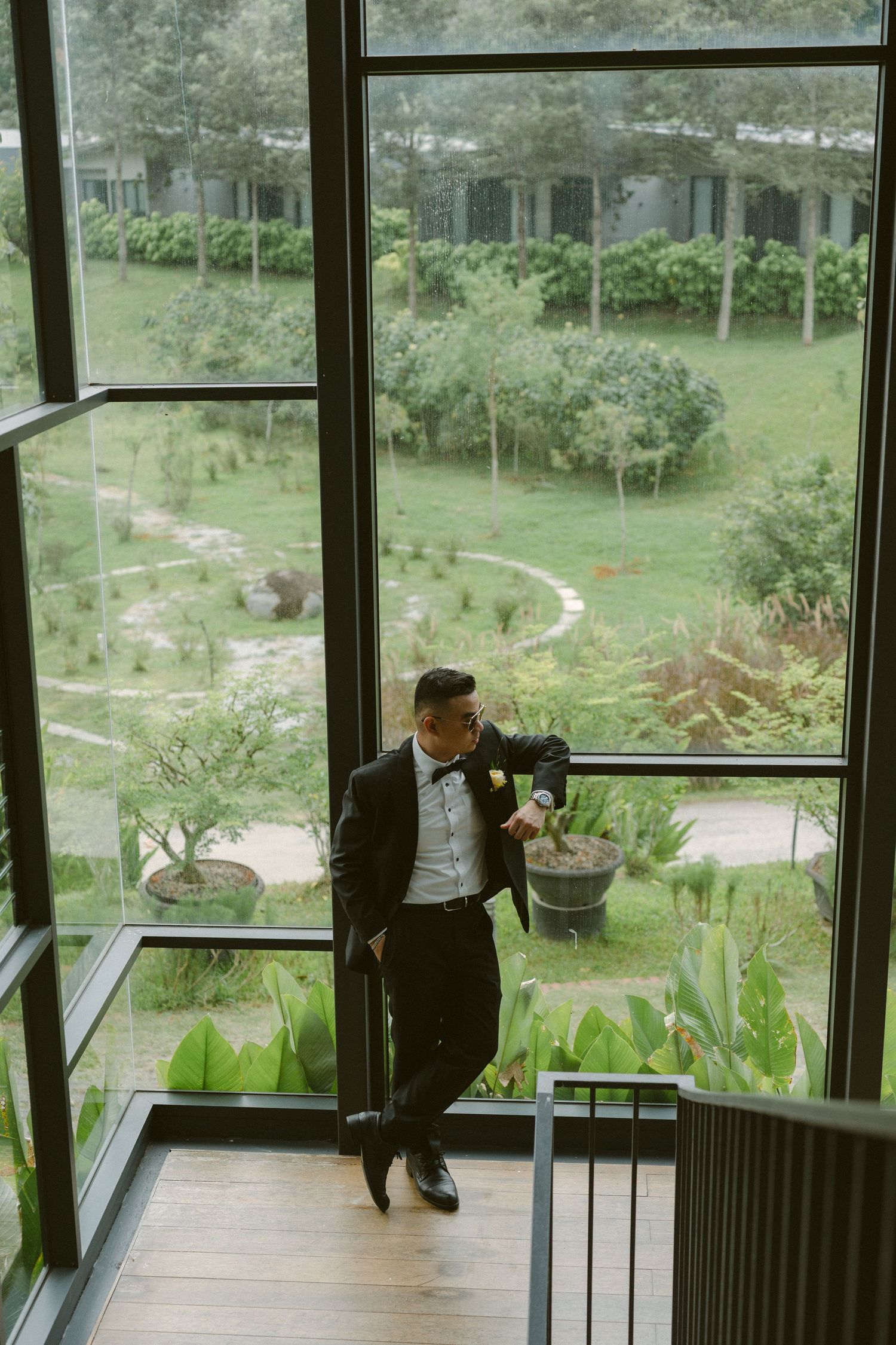 Person sitting on steps in a sunroom overlooking a lush green garden through large glass windows.