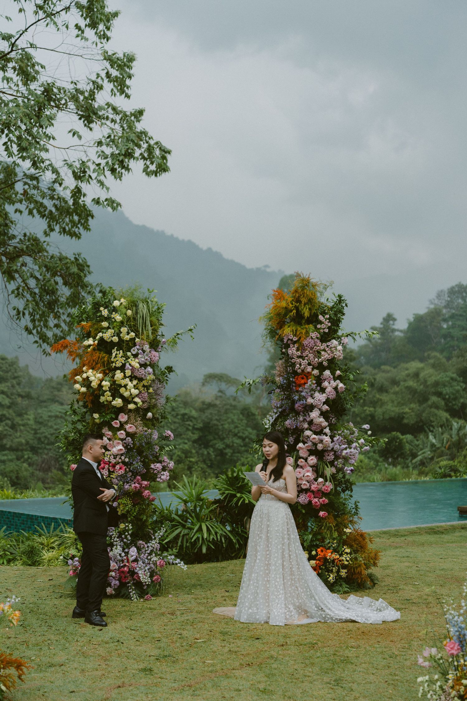 Wedding ceremony setup with floral arches by a scenic mountain lake, featuring lush decorations and misty landscape backdrop.