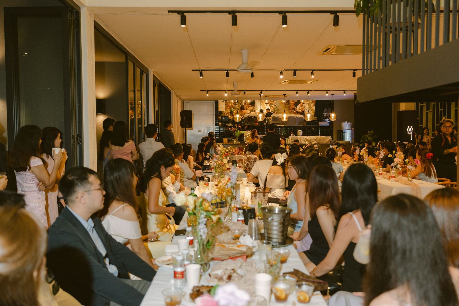 Wedding reception dinner party with guests seated at long tables in a warmly lit modern restaurant space.
