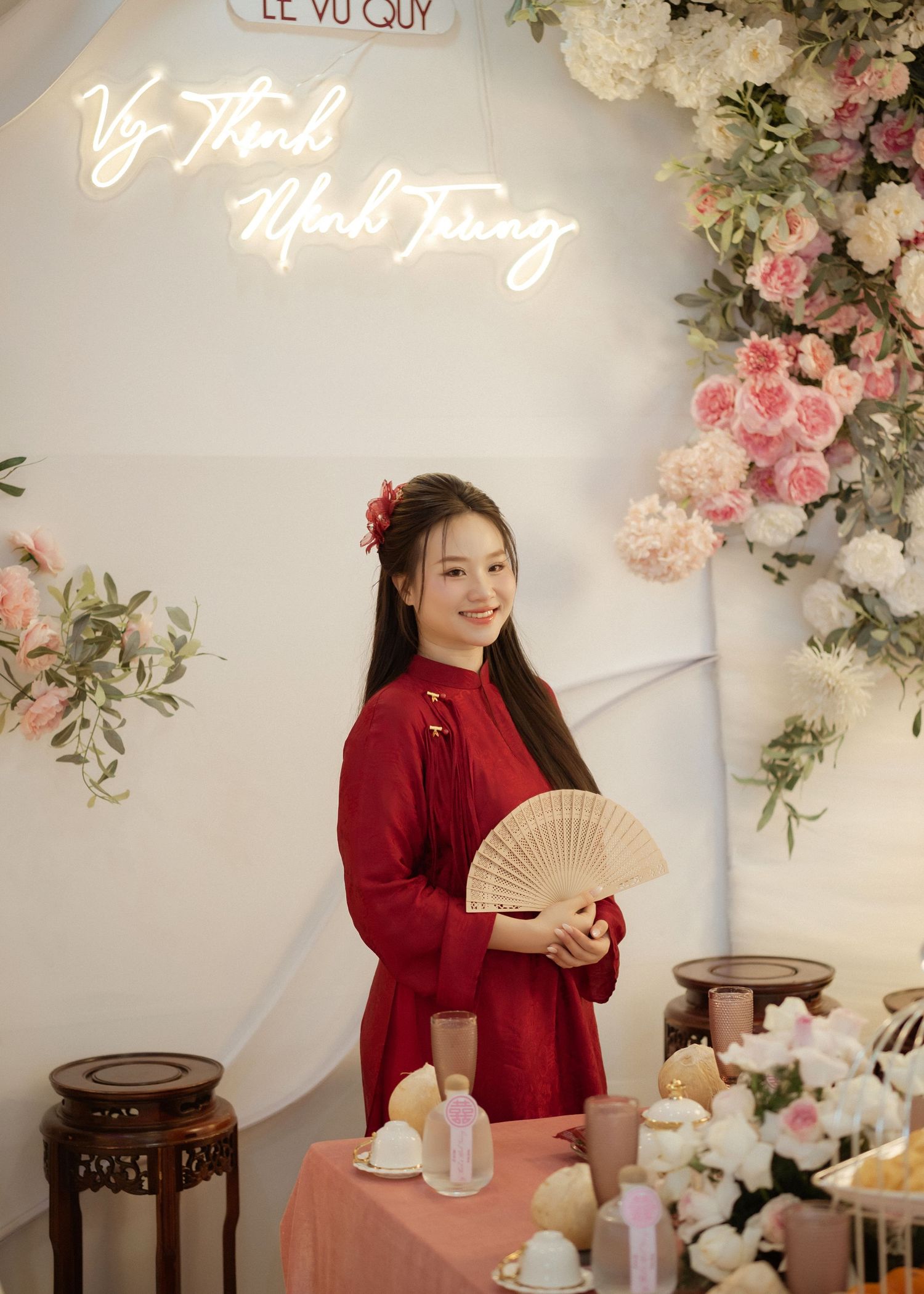 Person in a red traditional dress holding a folding fan poses under neon lights and floral decorations in an elegant setting.