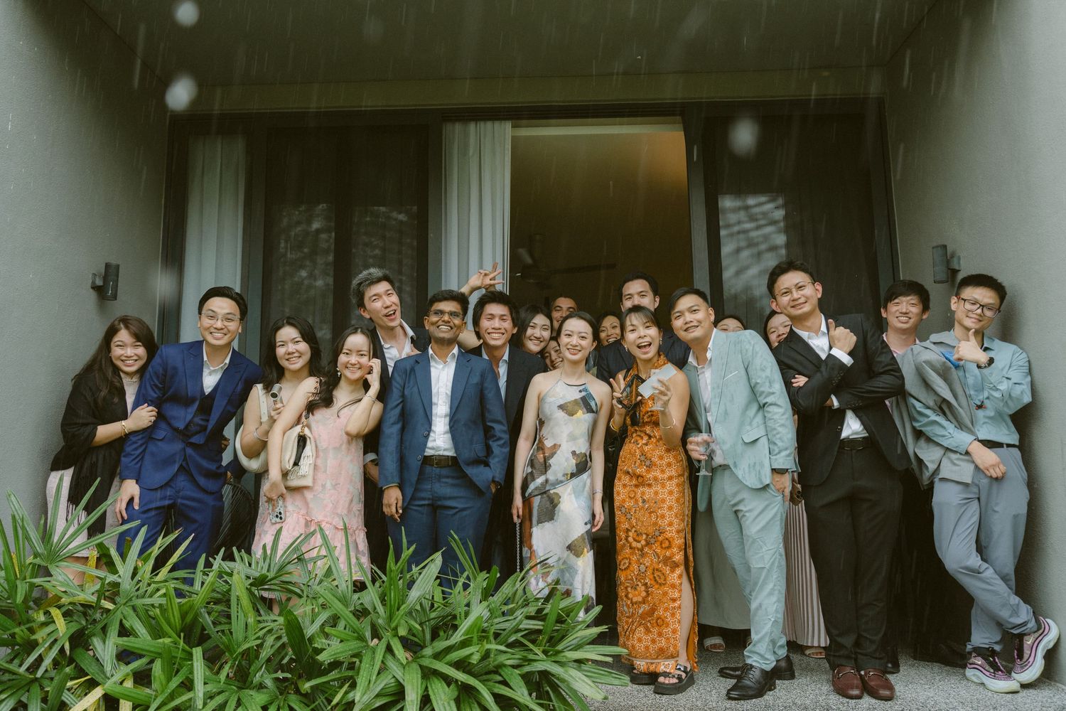 A group of guests pose for a photo outside a building entrance with greenery in the foreground.