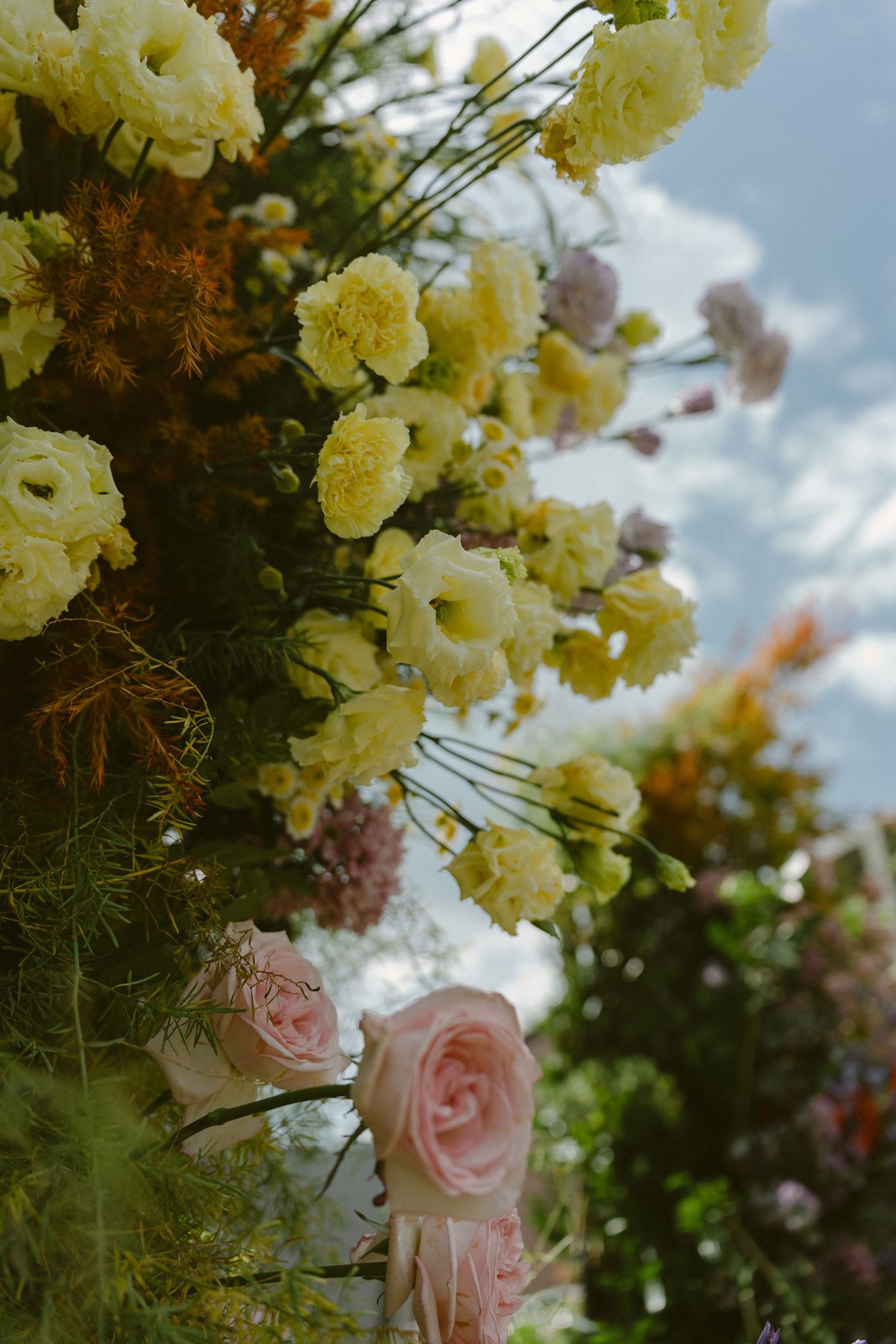 A garden arrangement featuring delicate pink roses and pale yellow flowers blooming against a soft blue sky backdrop.