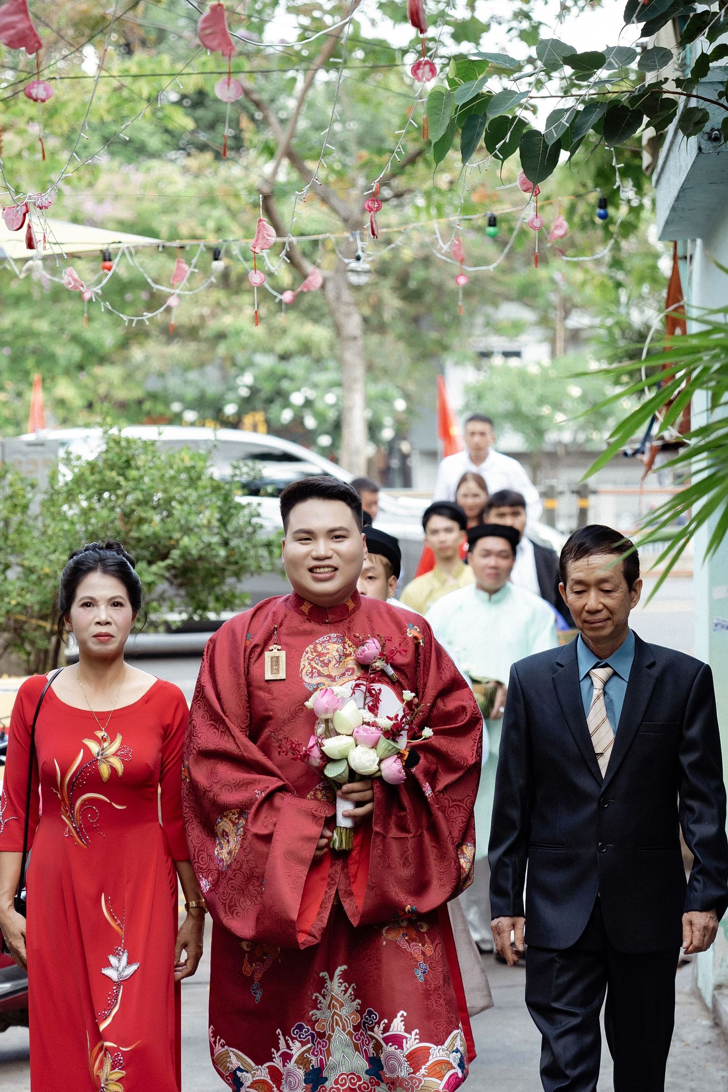 A wedding party walks outdoors with the groom wearing traditional red Asian formal attire holding flowers.